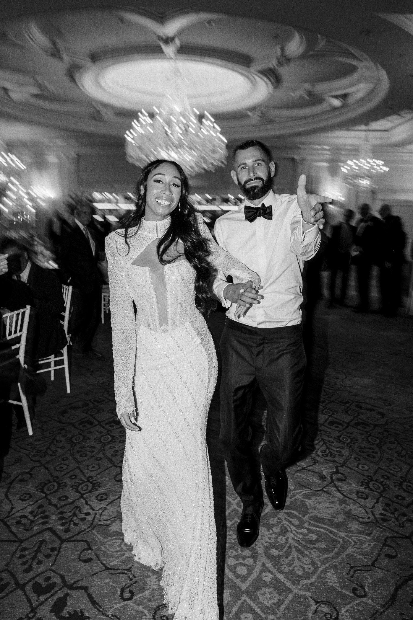A bride and groom dancing at their wedding reception in a grand ballroom with chandeliers, with guests seated at tables in the background.