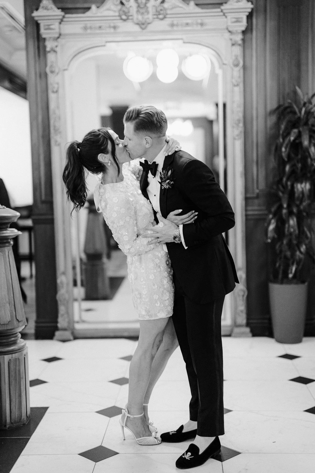 A black and white photo of a couple in wedding attire sharing a kiss inside a decorated room with a mirror and potted plants.