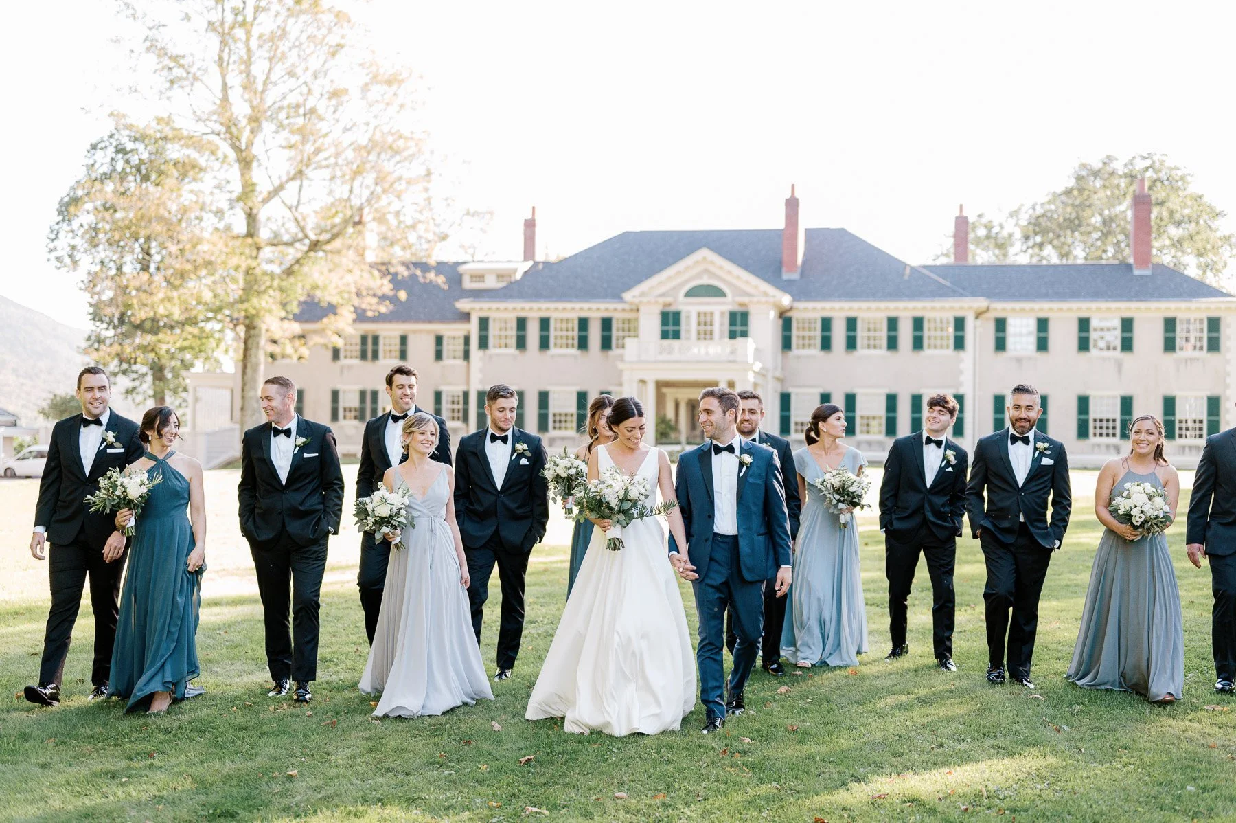 Wedding party walking on a lawn in front of a large white house with green shutters, the bride and groom in front, all dressed elegantly, holding bouquets, on a sunny day.