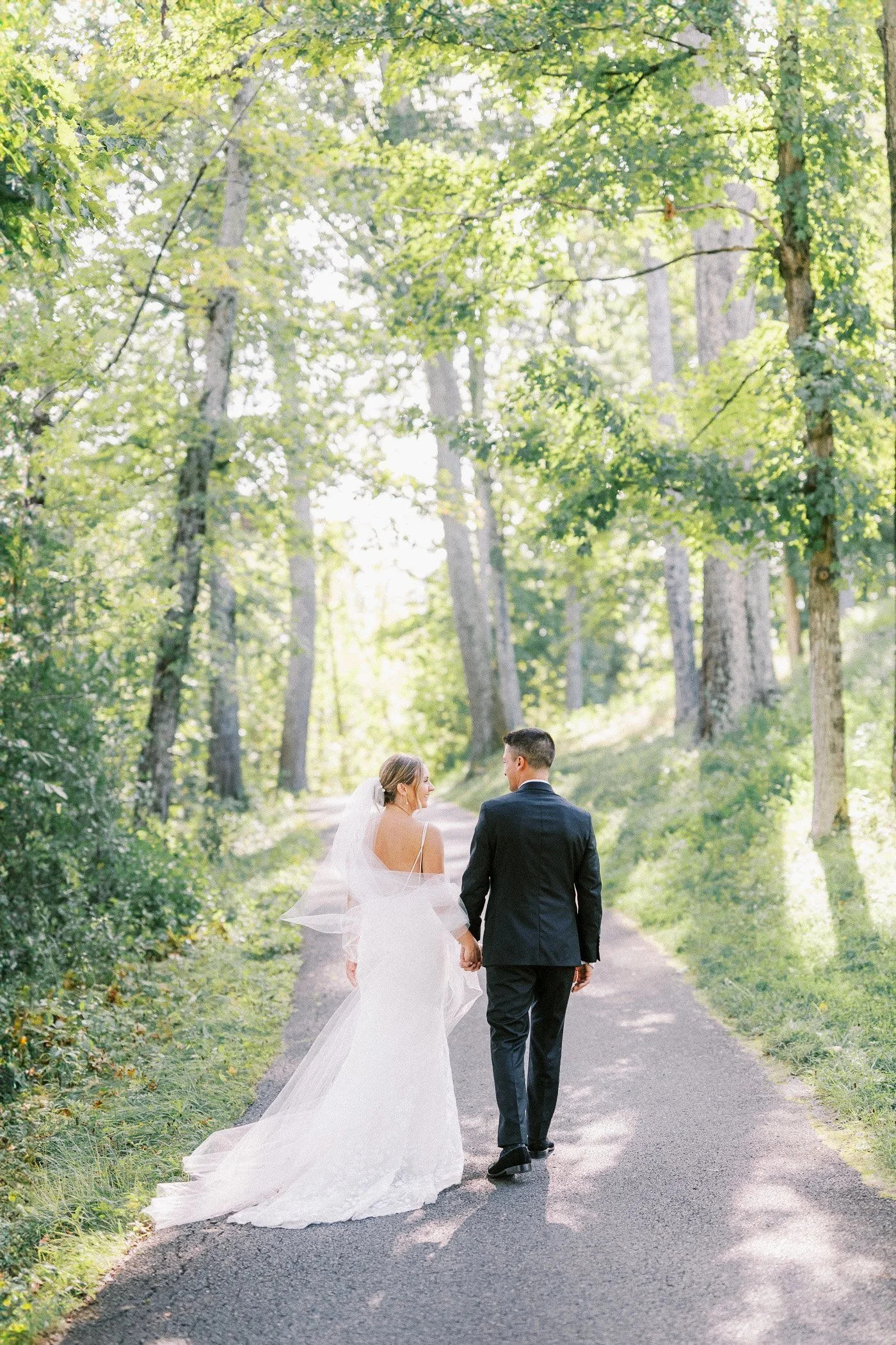 A bride and groom walking hand in hand down a forested country road, surrounded by tall green trees, on a sunny day.