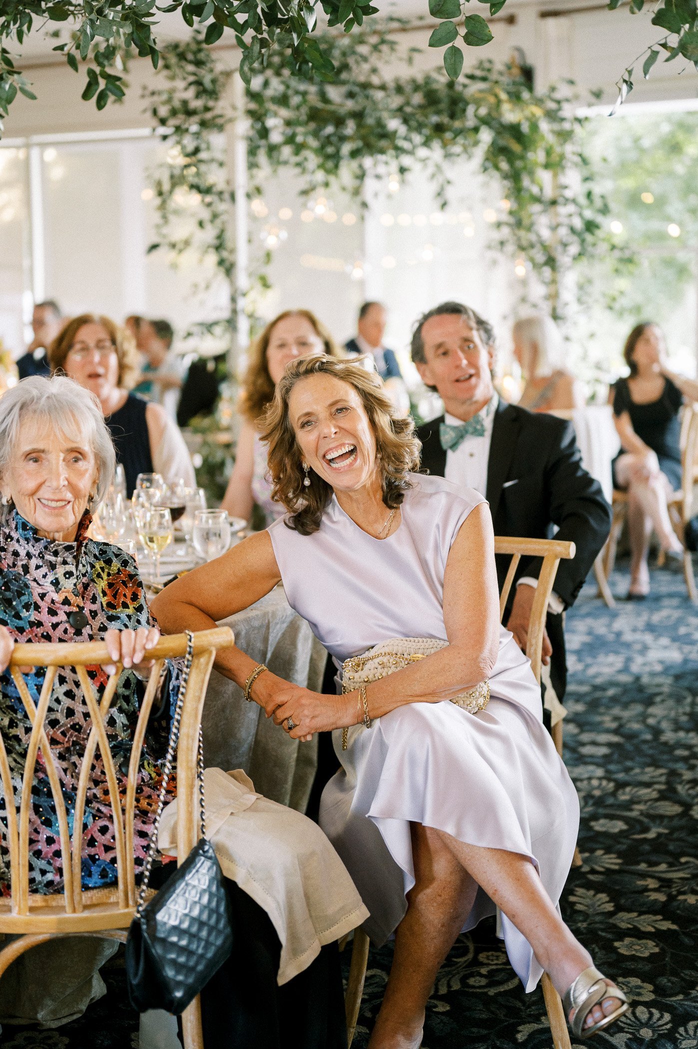A group of people at a formal event, seated at tables, smiling and laughing, with a woman in a silver dress in the foreground.