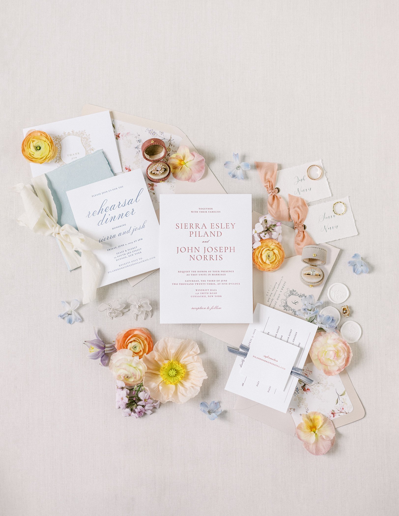 Wedding invitation suite with floral accents, rings, and small decorative elements on a light-colored surface.