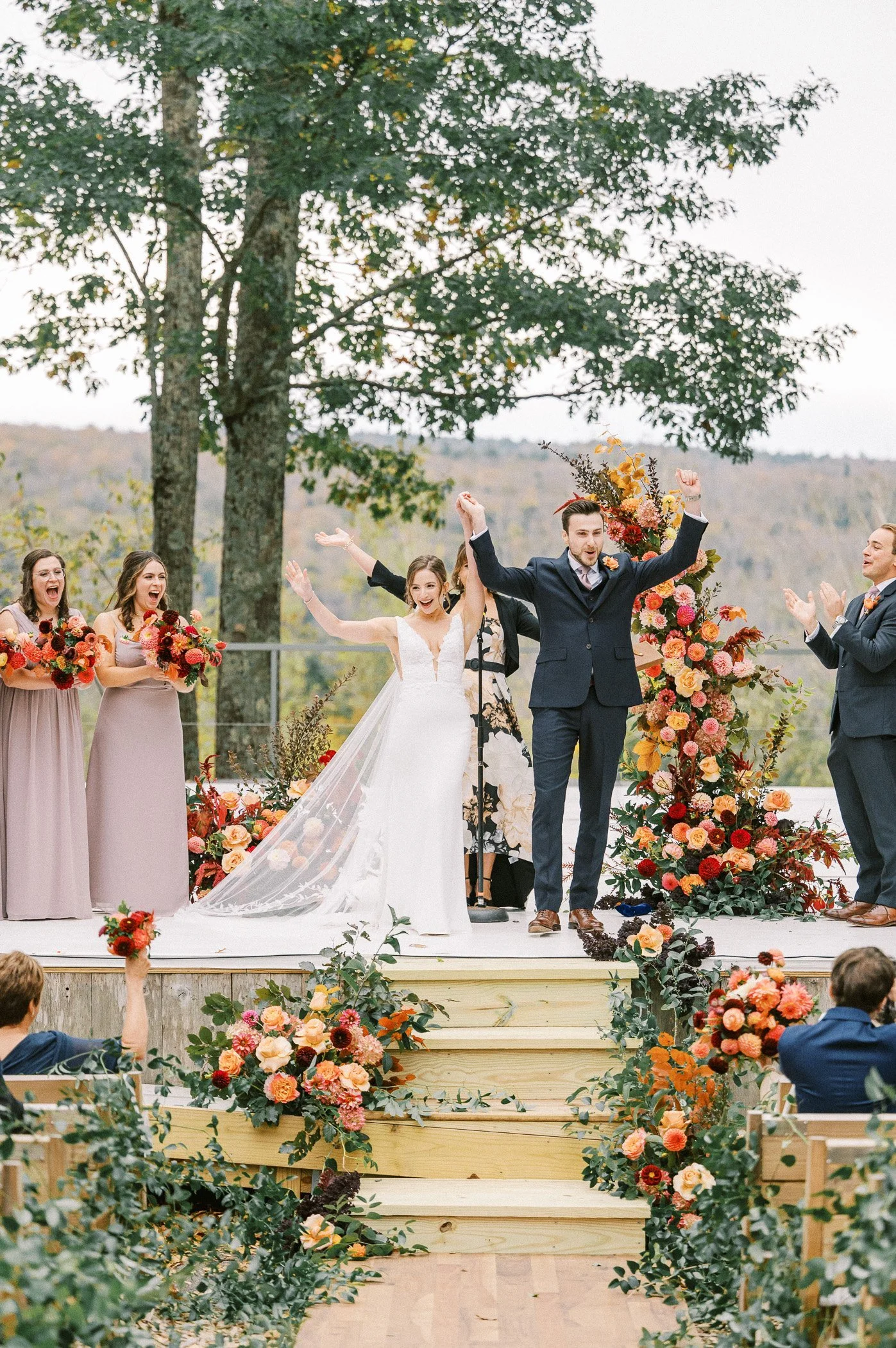 A wedding ceremony outdoors with the bride and groom celebrating on a decorated wooden deck, surrounded by bridesmaids, groomsmen, and floral arrangements, with scenic trees and hills in the background.