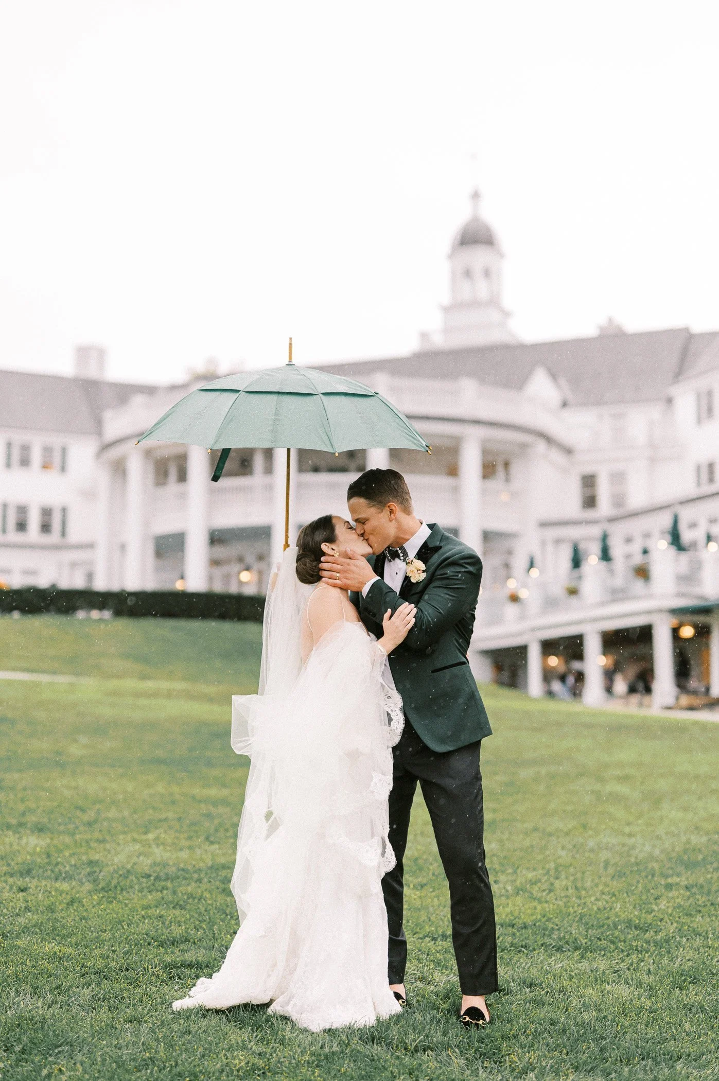 A bride and groom kissing under an umbrella on a rainy day outside a large white building, possibly a wedding venue.