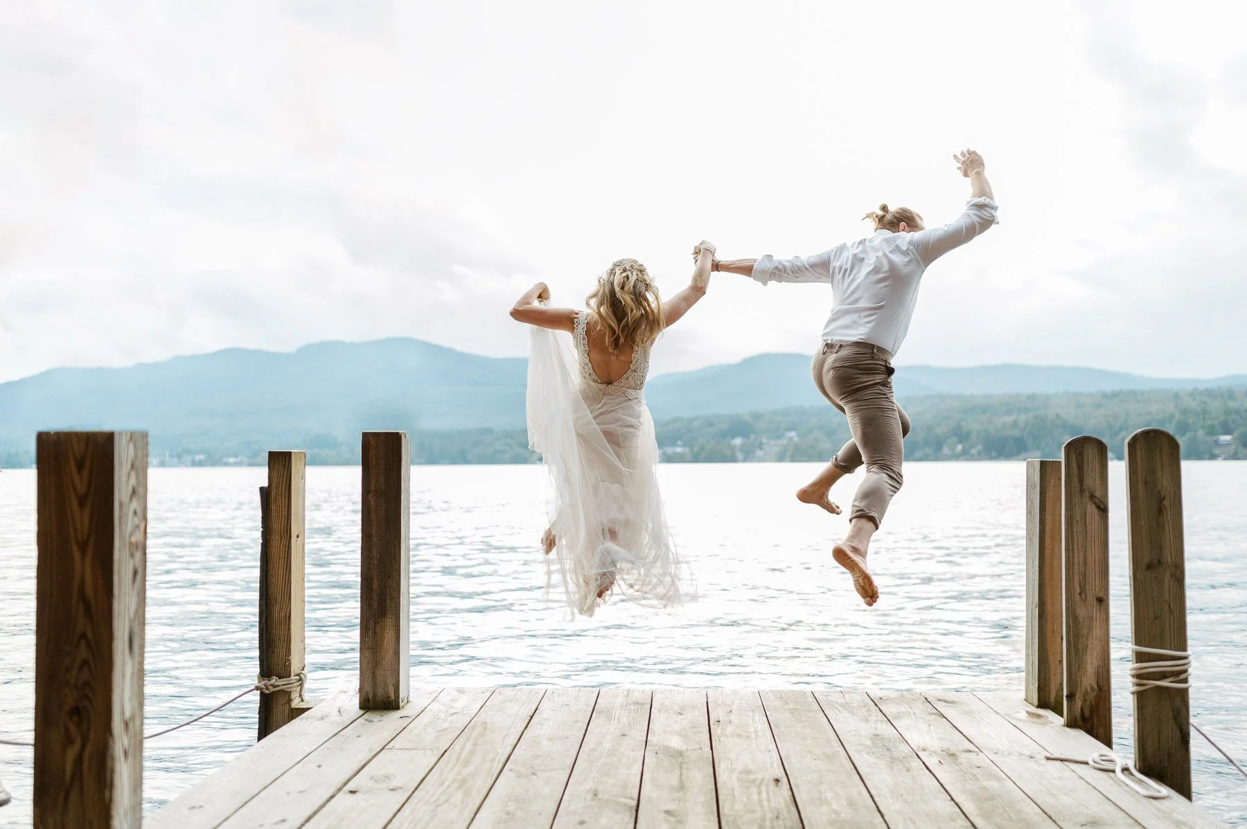 A couple jumping off a wooden dock into a lake with mountains in the background, smiling and holding hands.