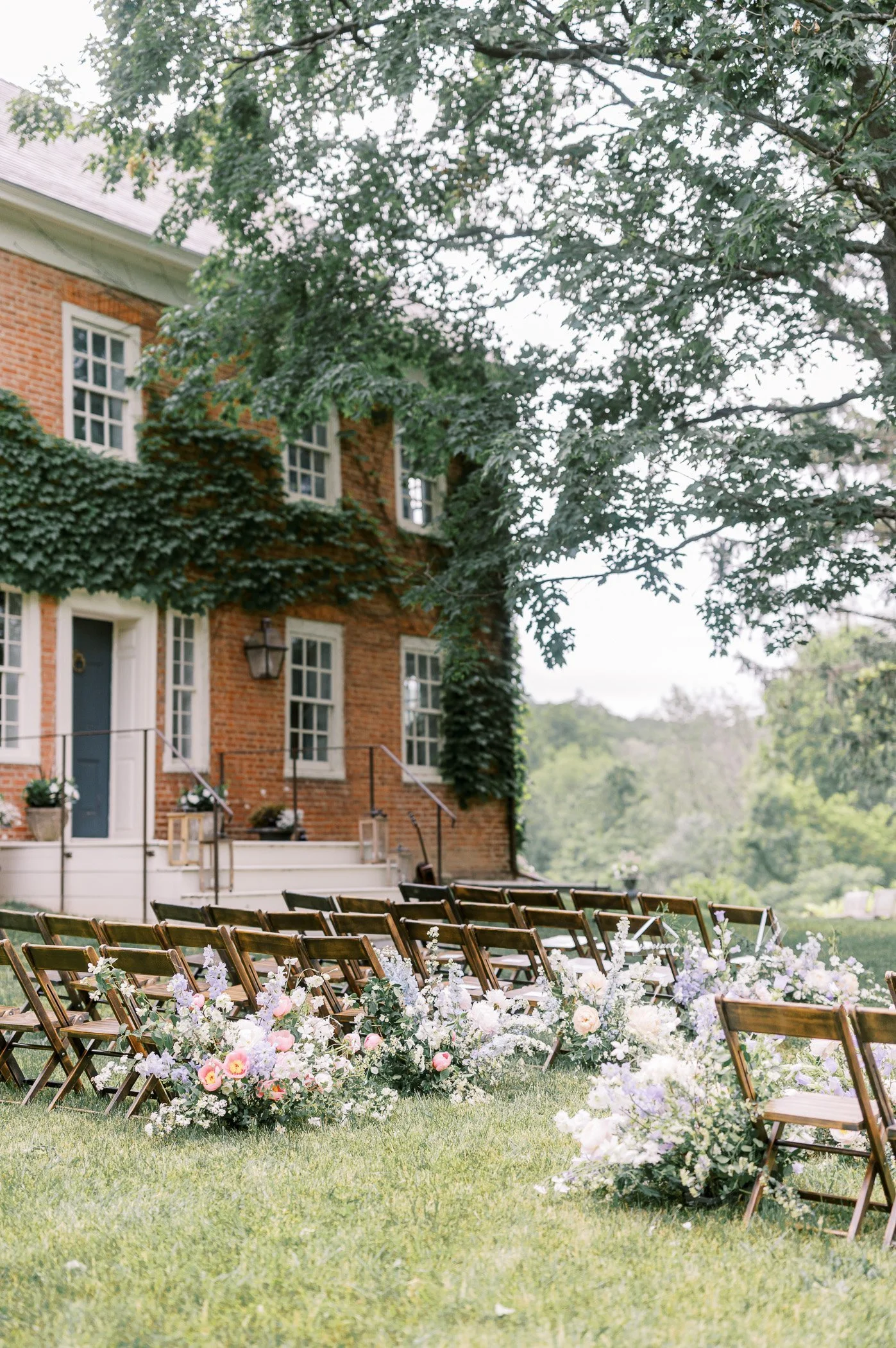 Outdoor wedding ceremony setup with wooden chairs and floral arrangements on green grass in front of a brick house with ivy-covered walls and large trees.