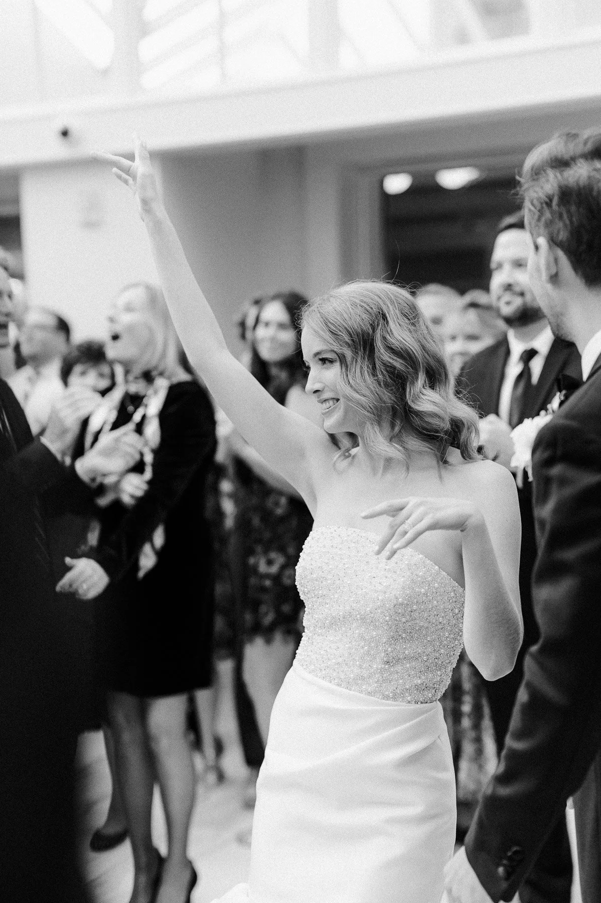 Black and white photo of a woman in a strapless dress celebrating at a formal event, surrounded by smiling people.