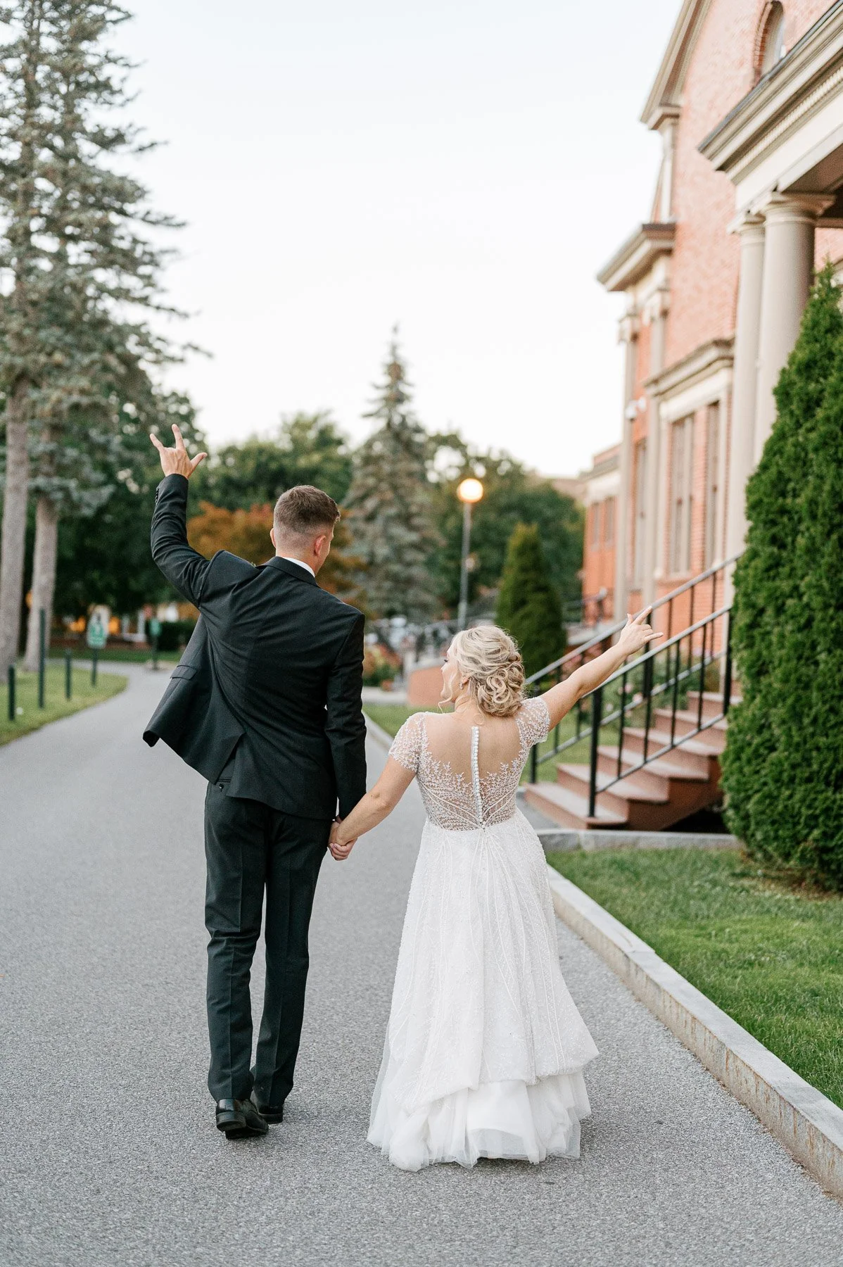 Bride and groom walking hand-in-hand on a sidewalk, holding hands with the groom raising one arm in celebration, near a brick building and greenery.