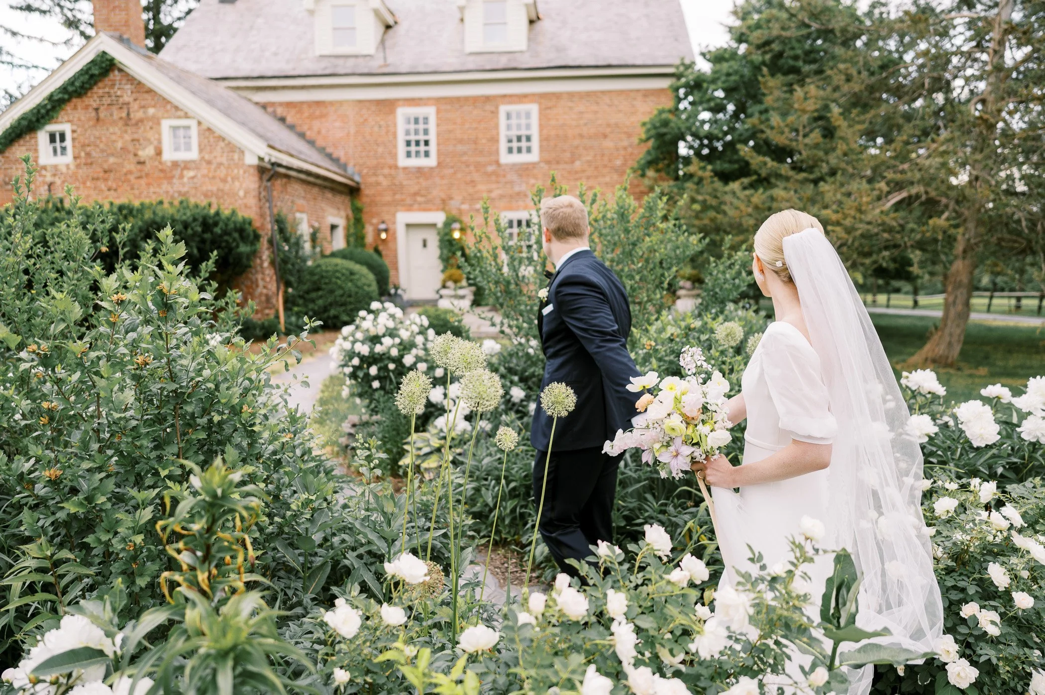 A bride in a white dress and veil holds a bouquet of flowers, walking in a garden with a groom in a dark suit, near a brick house with greenery and blooming flowers.