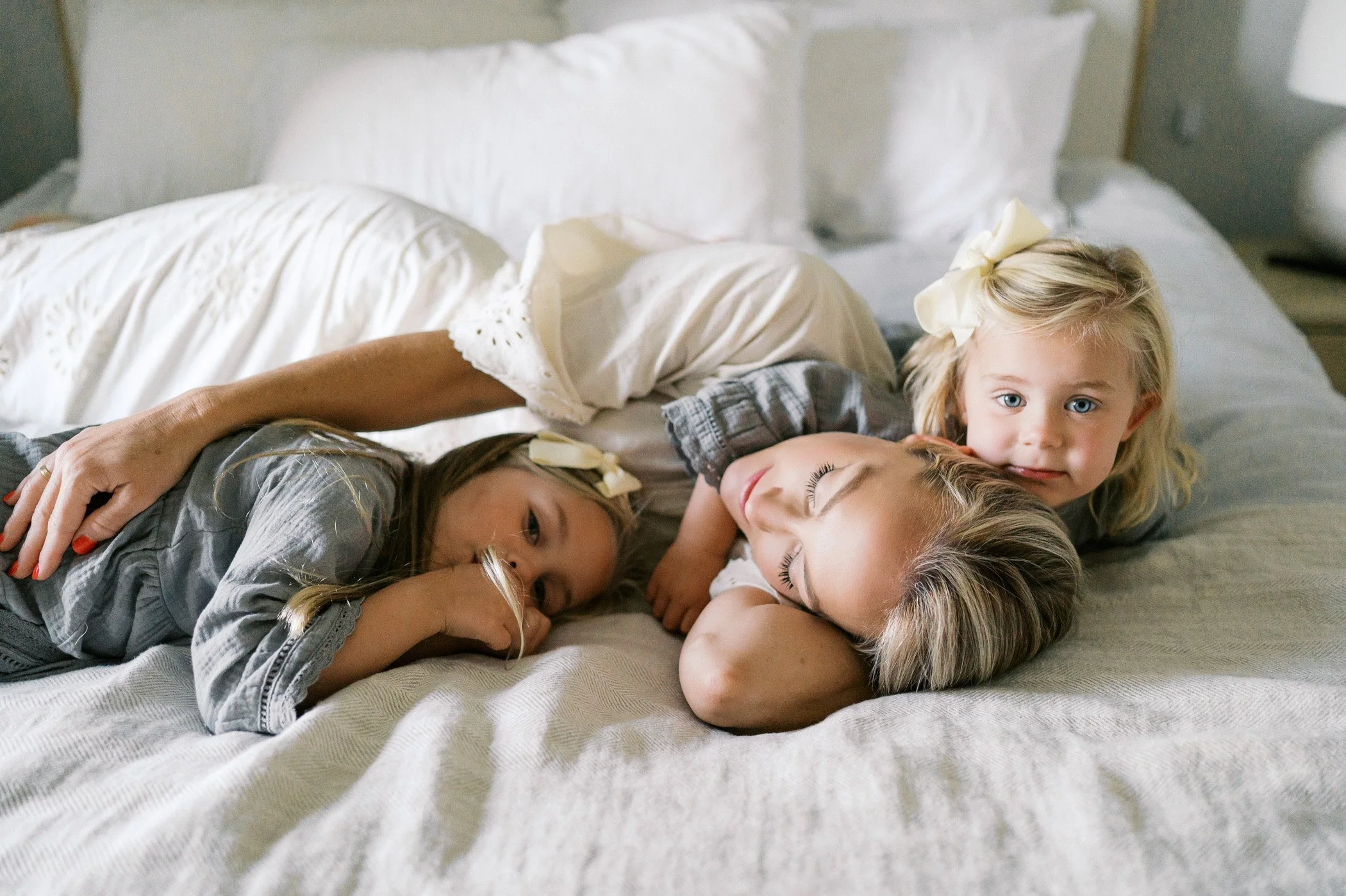 Two young girls lying on a bed, one with her eyes closed and the other looking at the camera, in a cozy bedroom setting.