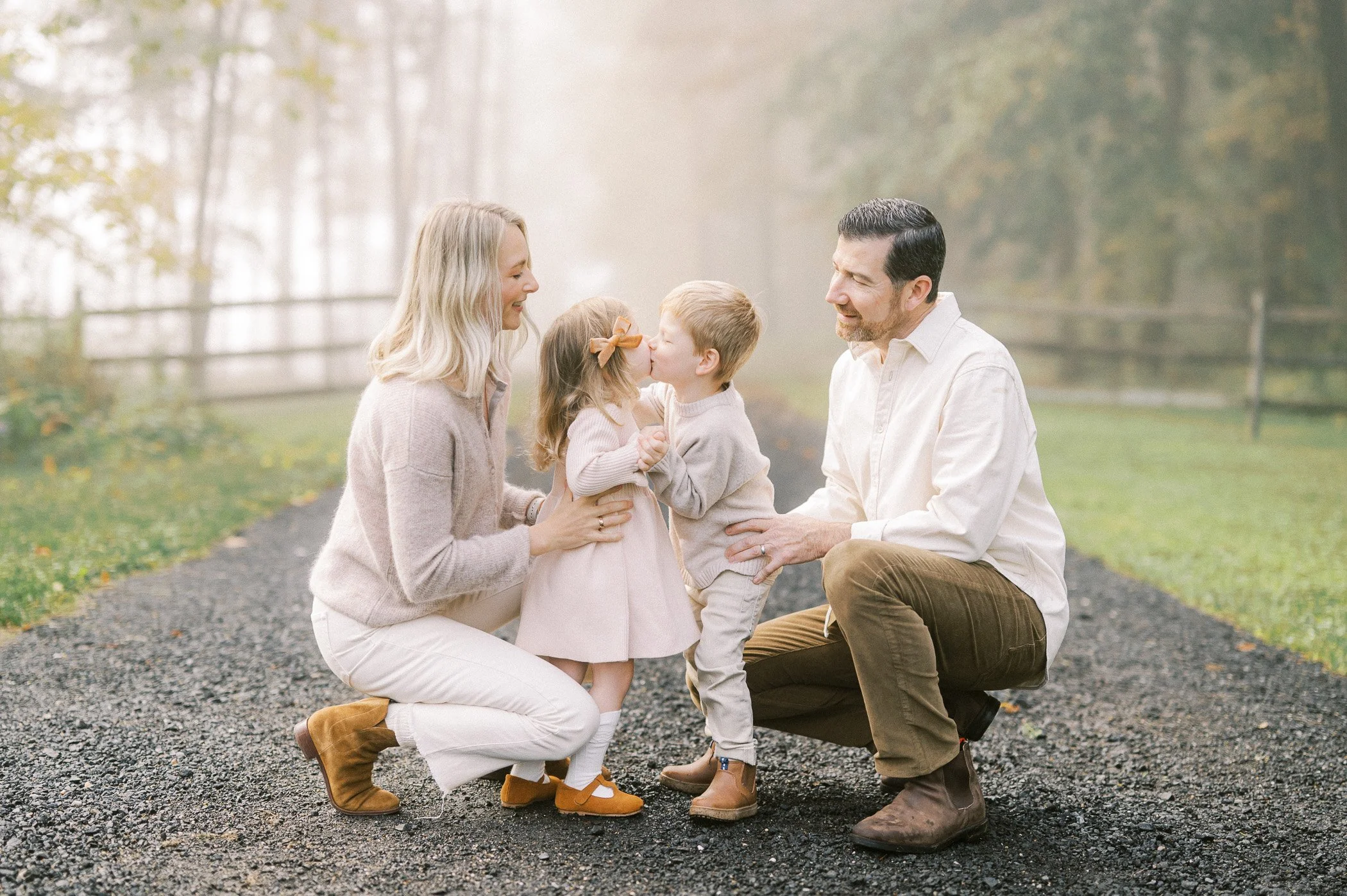 A family of four on a foggy outdoor path, two children giving each other a kiss while parents look on happily.