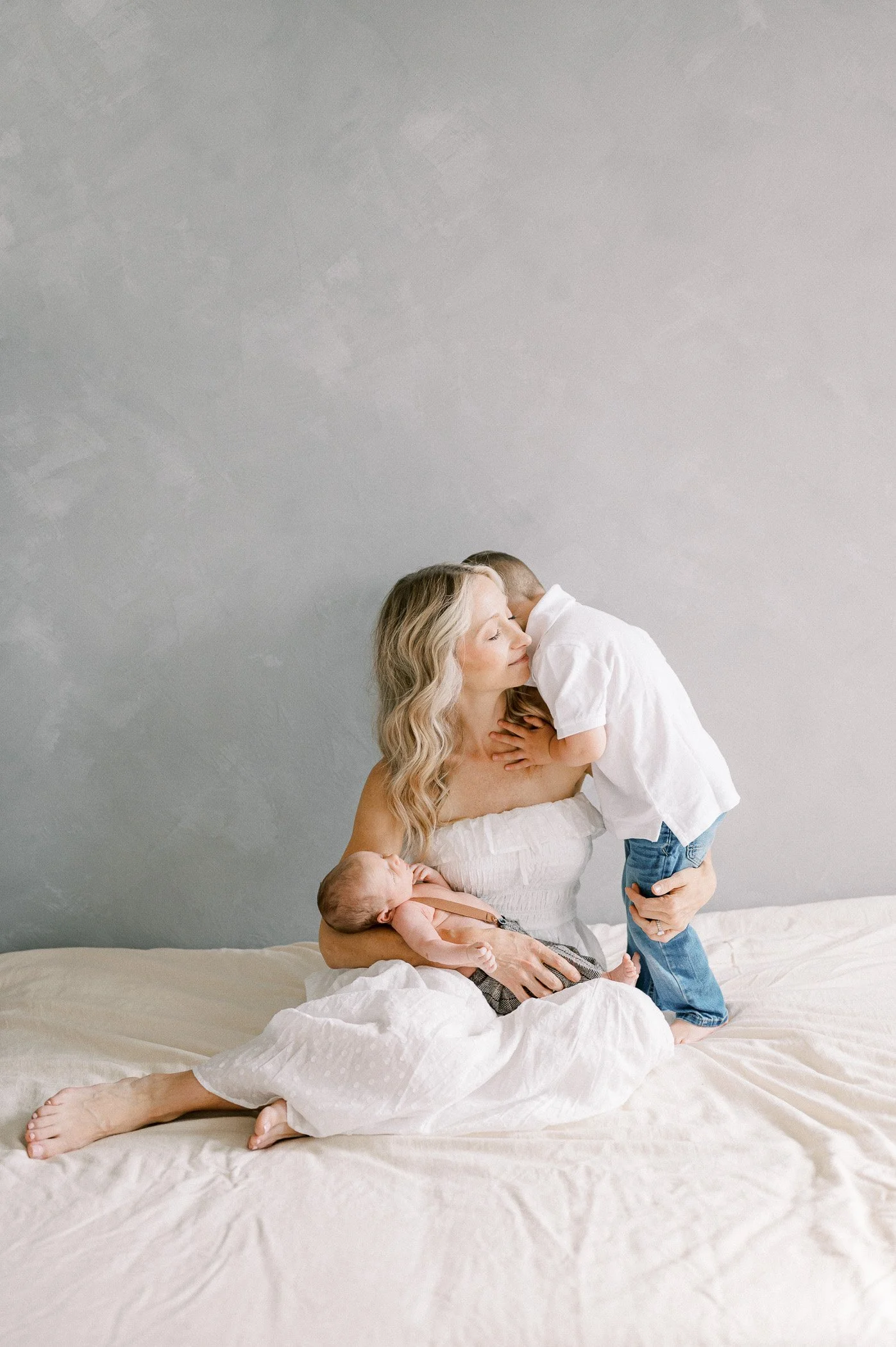 A woman with wavy blonde hair sitting on a bed, holding a newborn in her lap while a young boy kisses her on the cheek.