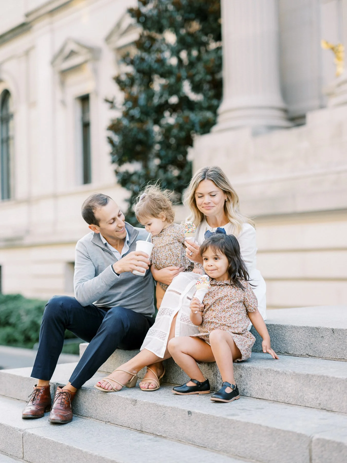 Family of four sitting on steps outside a building, enjoying ice cream and drinks, with the parents smiling and the two young girls eating ice cream.