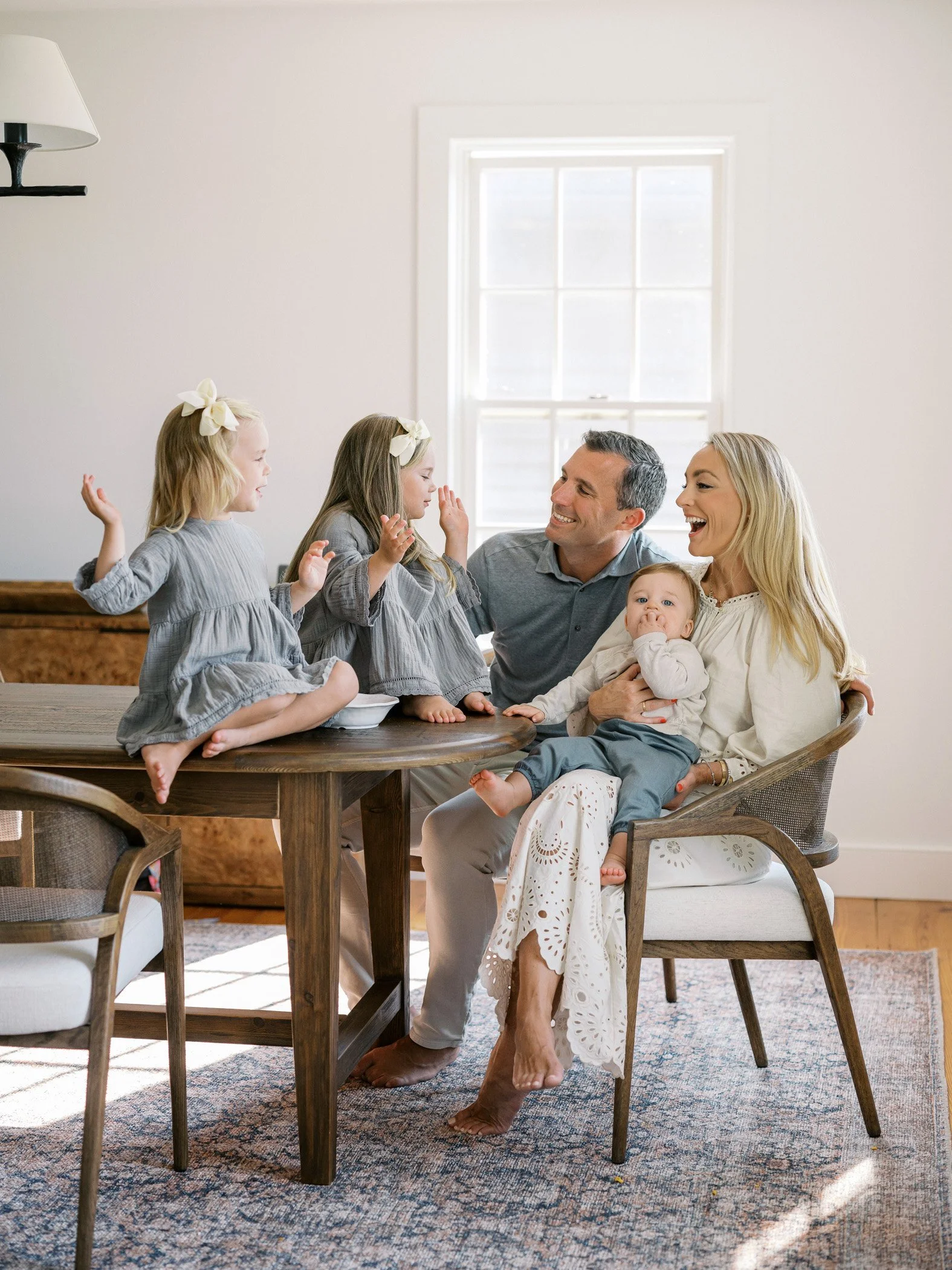 Family of six sitting around a dining table, smiling and enjoying time together in a bright room with a window in the background.