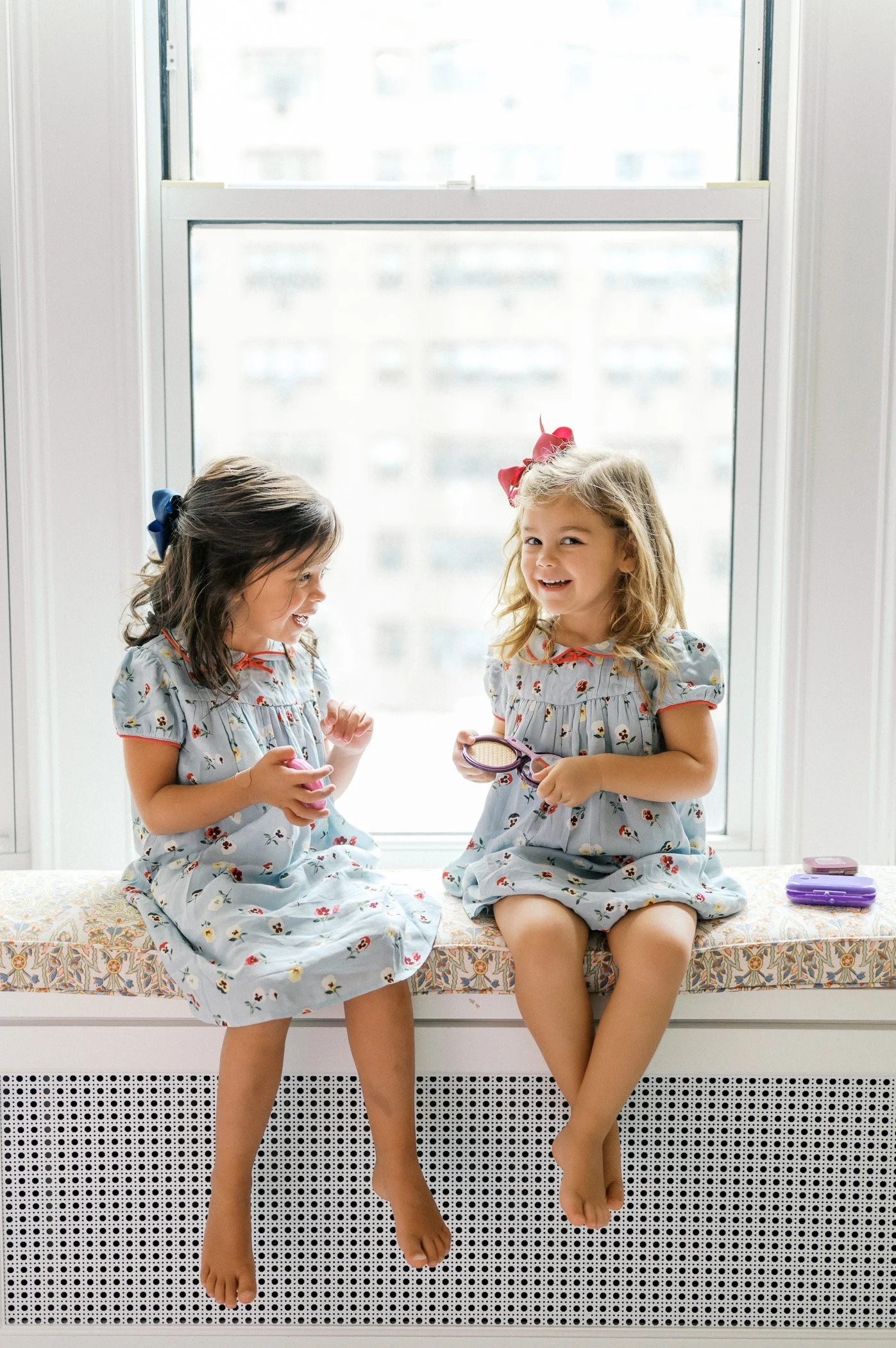 Two young girls sitting on a windowsill, playing with toy makeup, smiling and laughing, wearing matching floral dresses with bows in their hair.