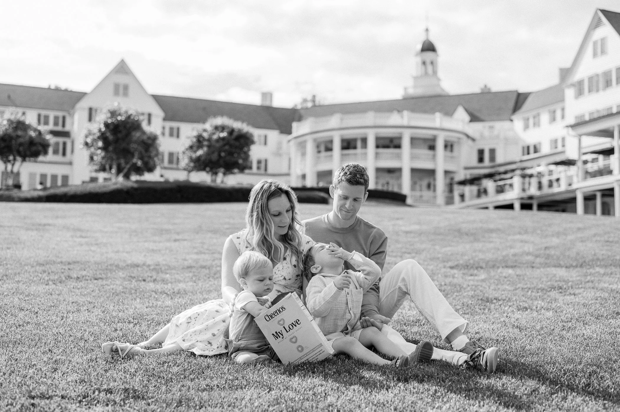 A family of four sitting on a lawn, with a large house in the background. The mother and father are with their two young children, a boy and a girl. The girl is holding a box of Cheerios, and the boy appears to be eating or touching his face.