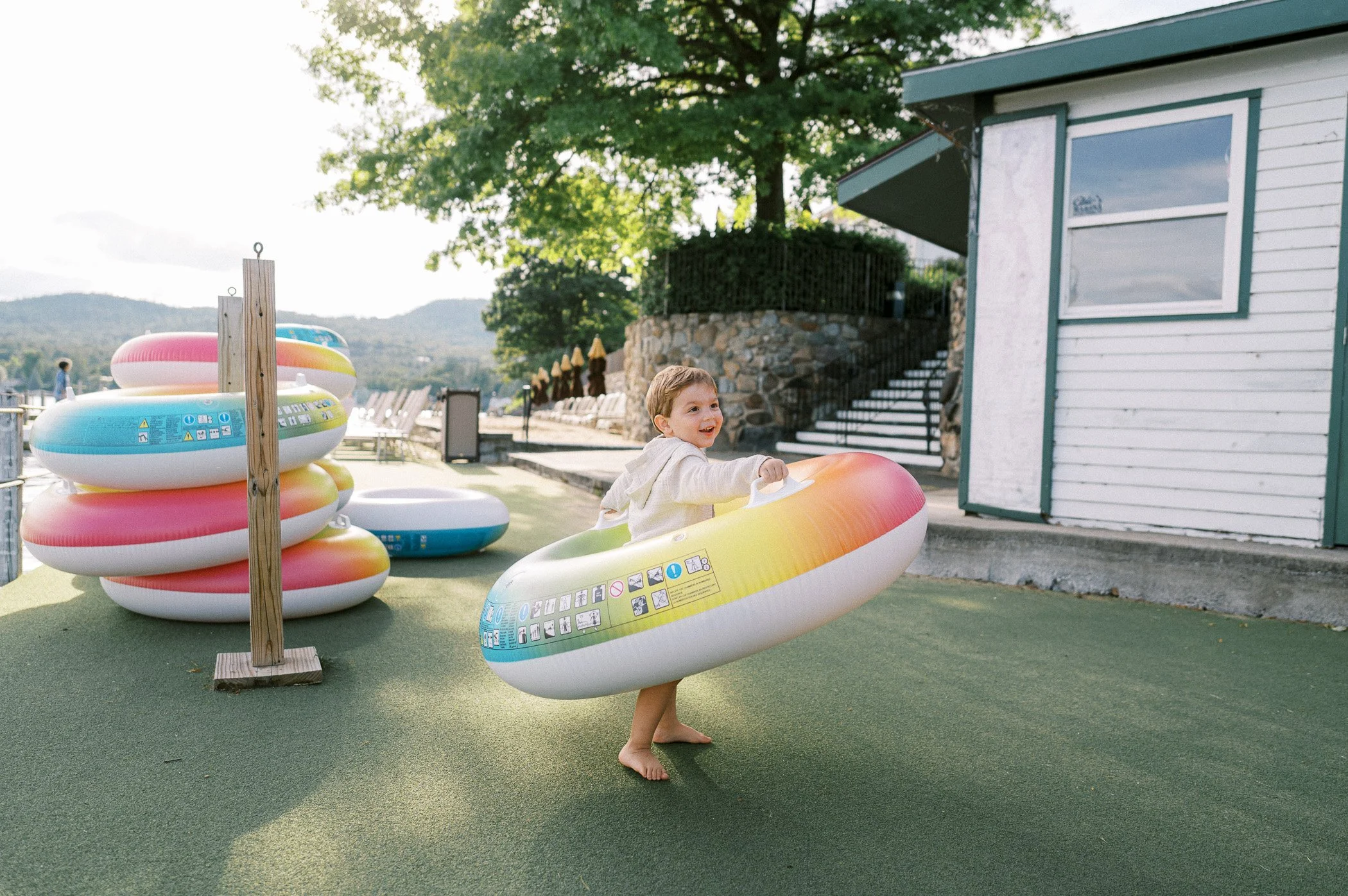 A young child smiling and playing in a large inflatable pool ring outdoors on a sunny day at a water park or pool area, with other pool rings stacked nearby, a building and stairs in the background, and lush green trees and mountains in the distance.