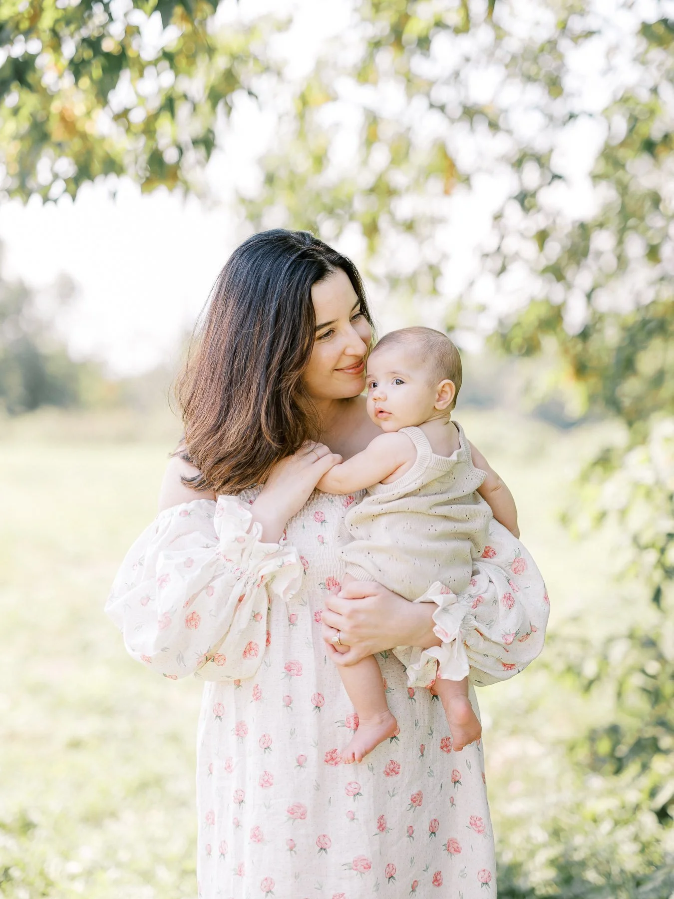 A woman with dark hair holding a baby girl outdoors on a sunny day, with trees and greenery in the background.