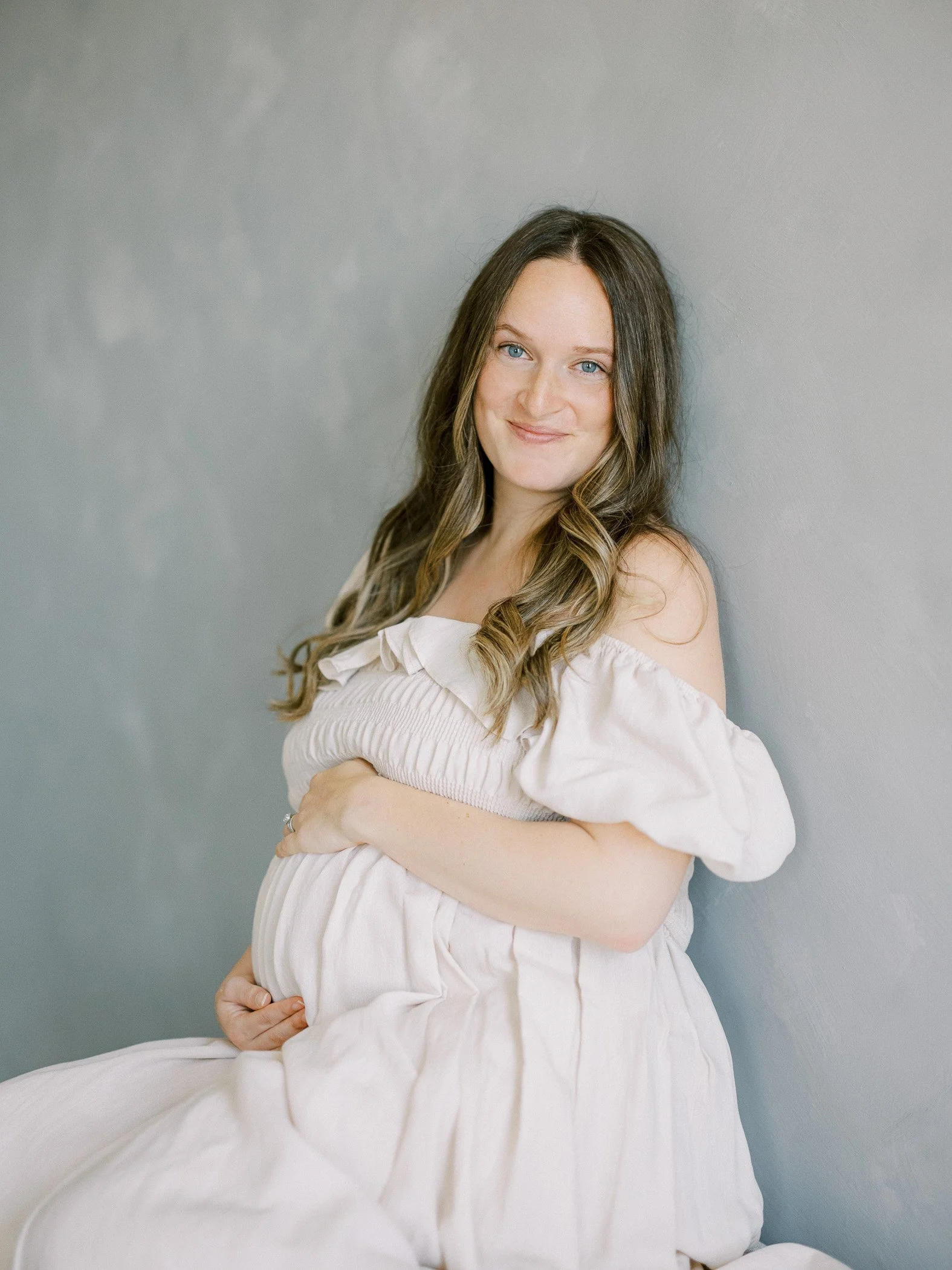 A pregnant woman with long brown hair and blue eyes, smiling softly, sitting against a plain gray wall, wearing an off-white dress with puffed sleeves.