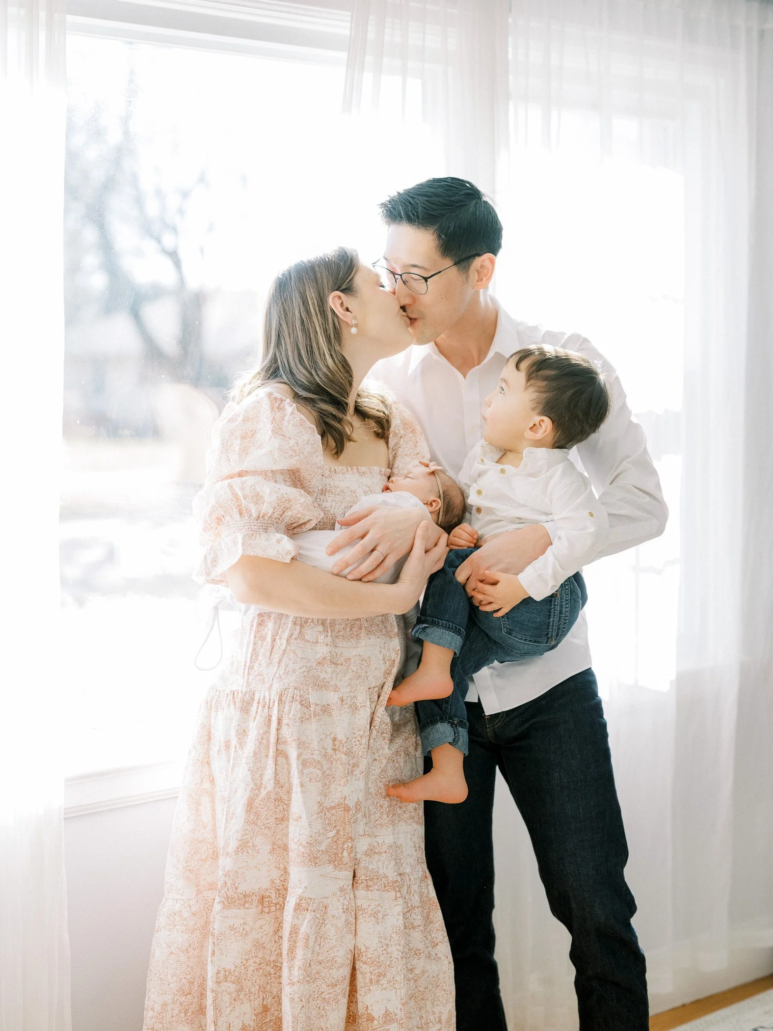 A family of four, including a woman, a man, a young boy, and an infant, sharing a tender moment and a kiss in front of a bright window.