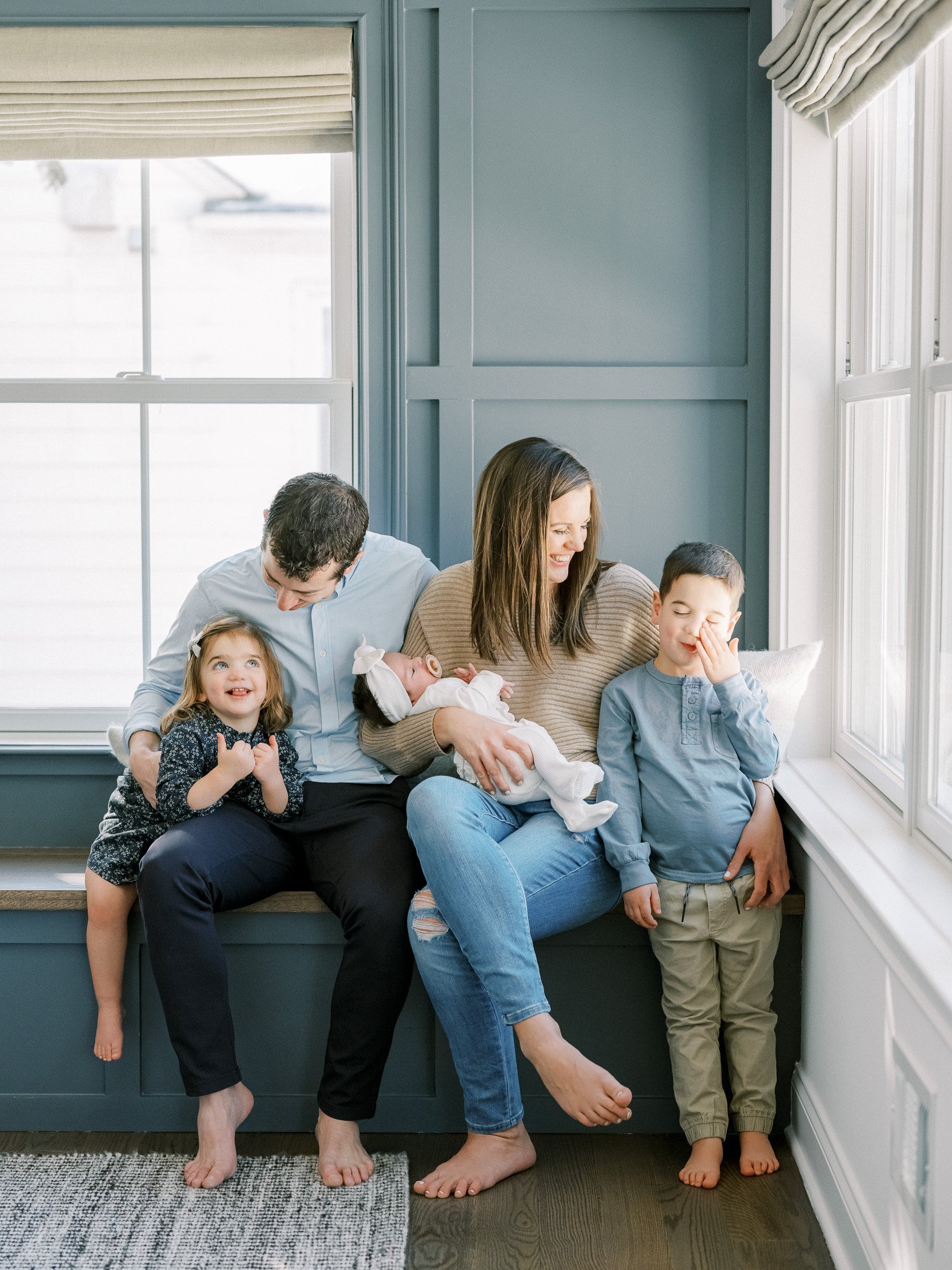 A family of five sitting on a window seat in a bright room, smiling and enjoying each other's company.