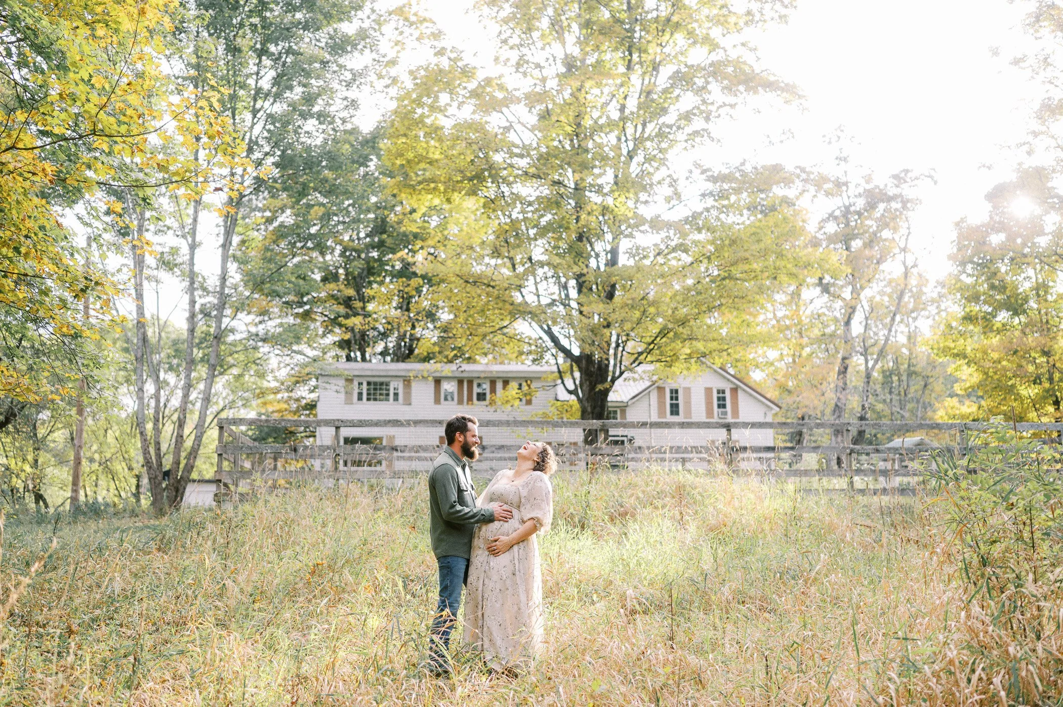 A couple standing outdoors in a field with tall grass, holding hands and smiling, with a house and trees in the background under bright sunlight.