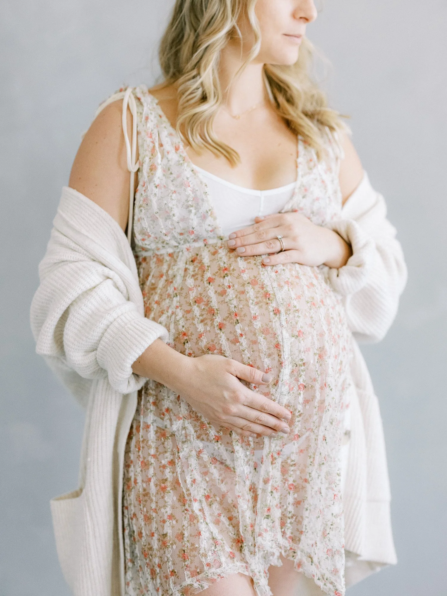 A pregnant woman wearing a floral dress and a white top, with her hands gently resting on her belly.