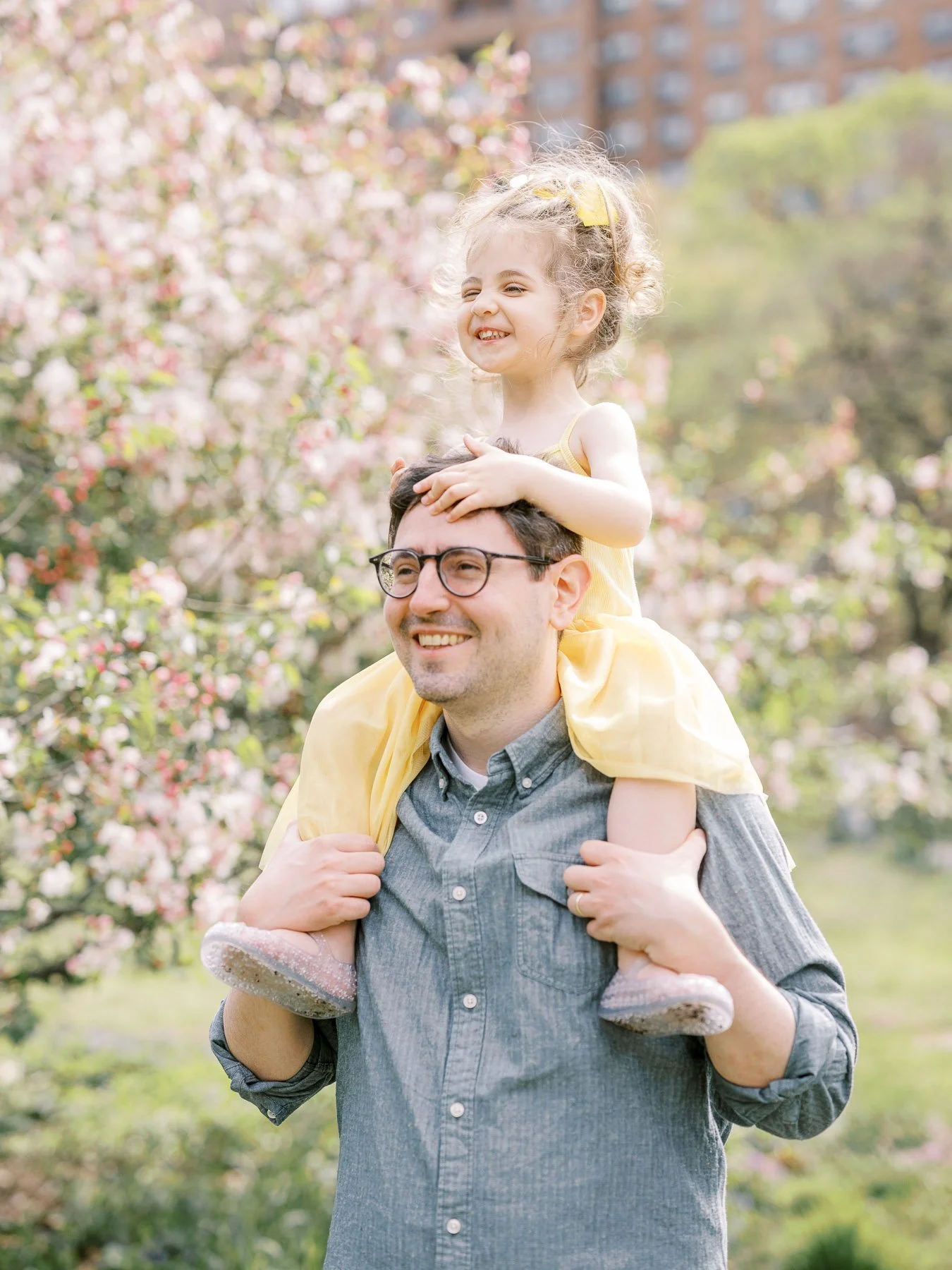 A man with glasses carrying a young girl on his shoulders in a park with blooming trees, both smiling and enjoying the outdoors.
