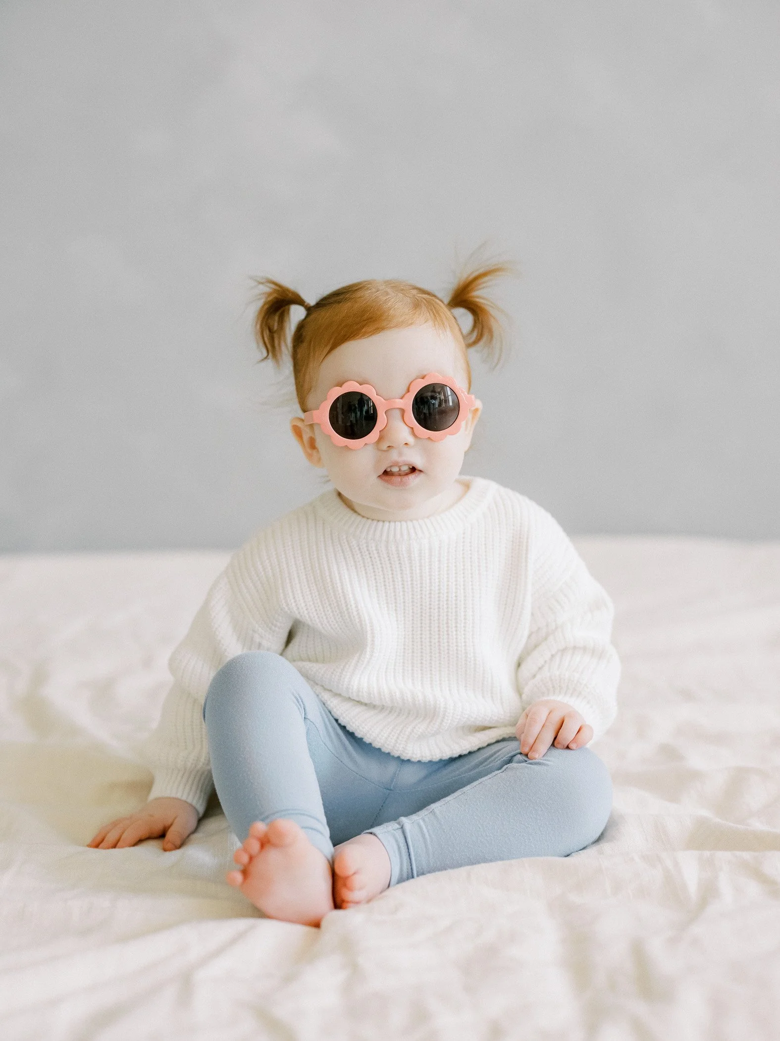 A young girl with red hair styled in pigtails, wearing pink sunglasses, a white sweater, and light blue leggings, sitting on a white bed.