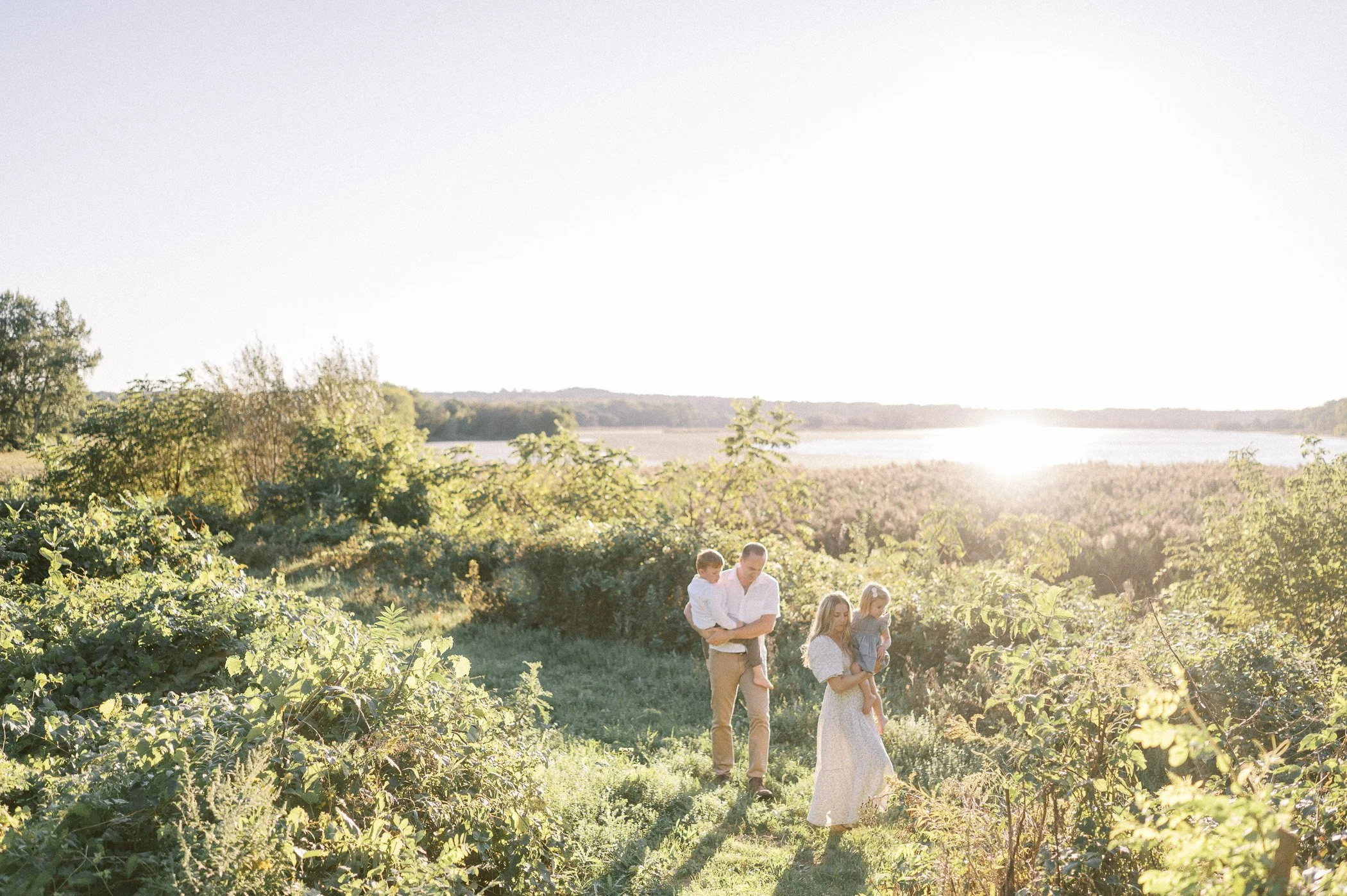 A family walking through a lush green field at sunset, with a lake in the background.