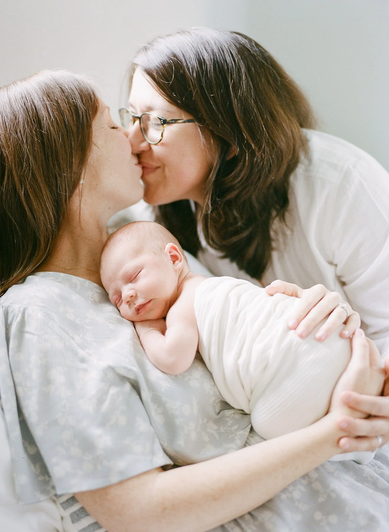 A woman and a woman kissing while holding a sleeping baby on her arm.