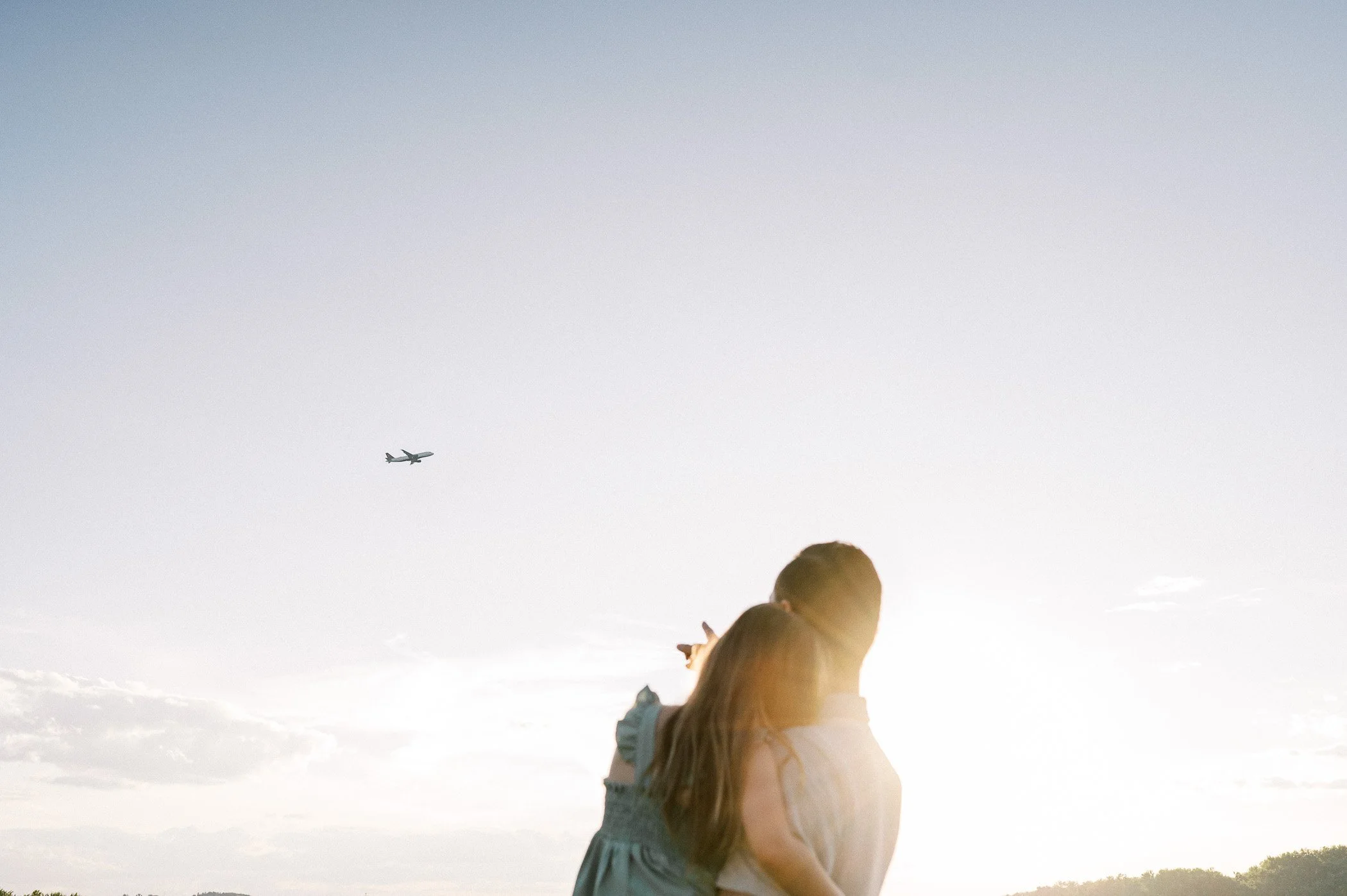 A woman and a young girl standing together outdoors during sunset, with the woman pointing towards an airplane flying in the sky.