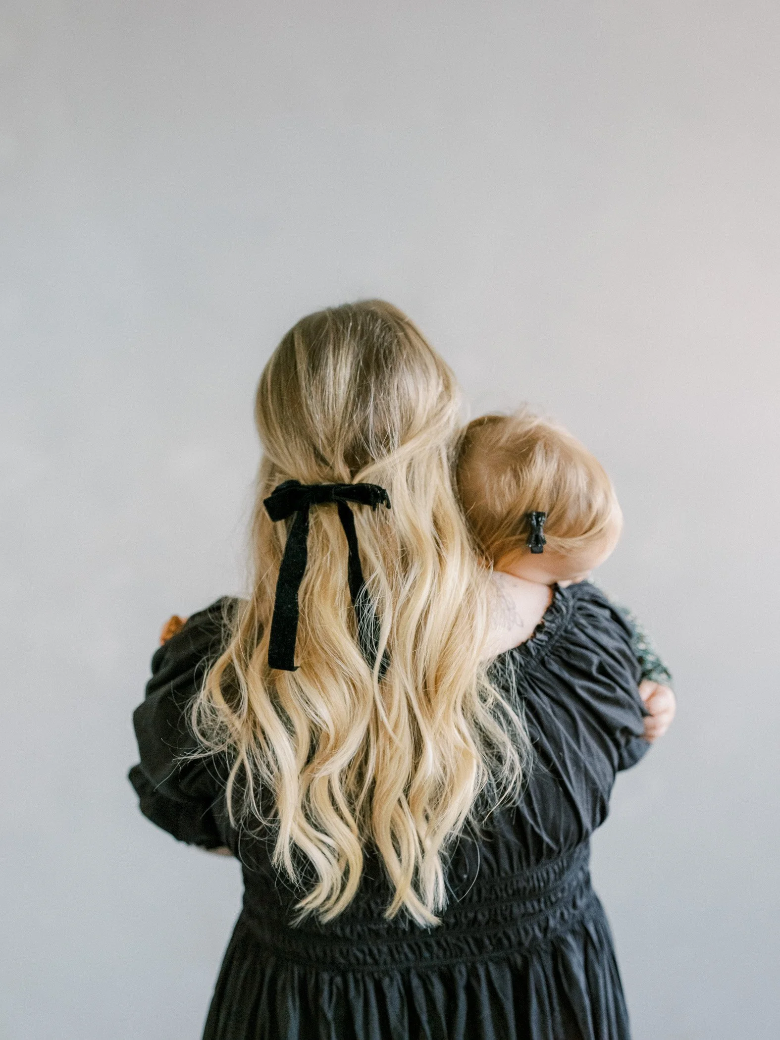 A woman with long, wavy blonde hair holding a small child with blonde hair against a plain gray background.