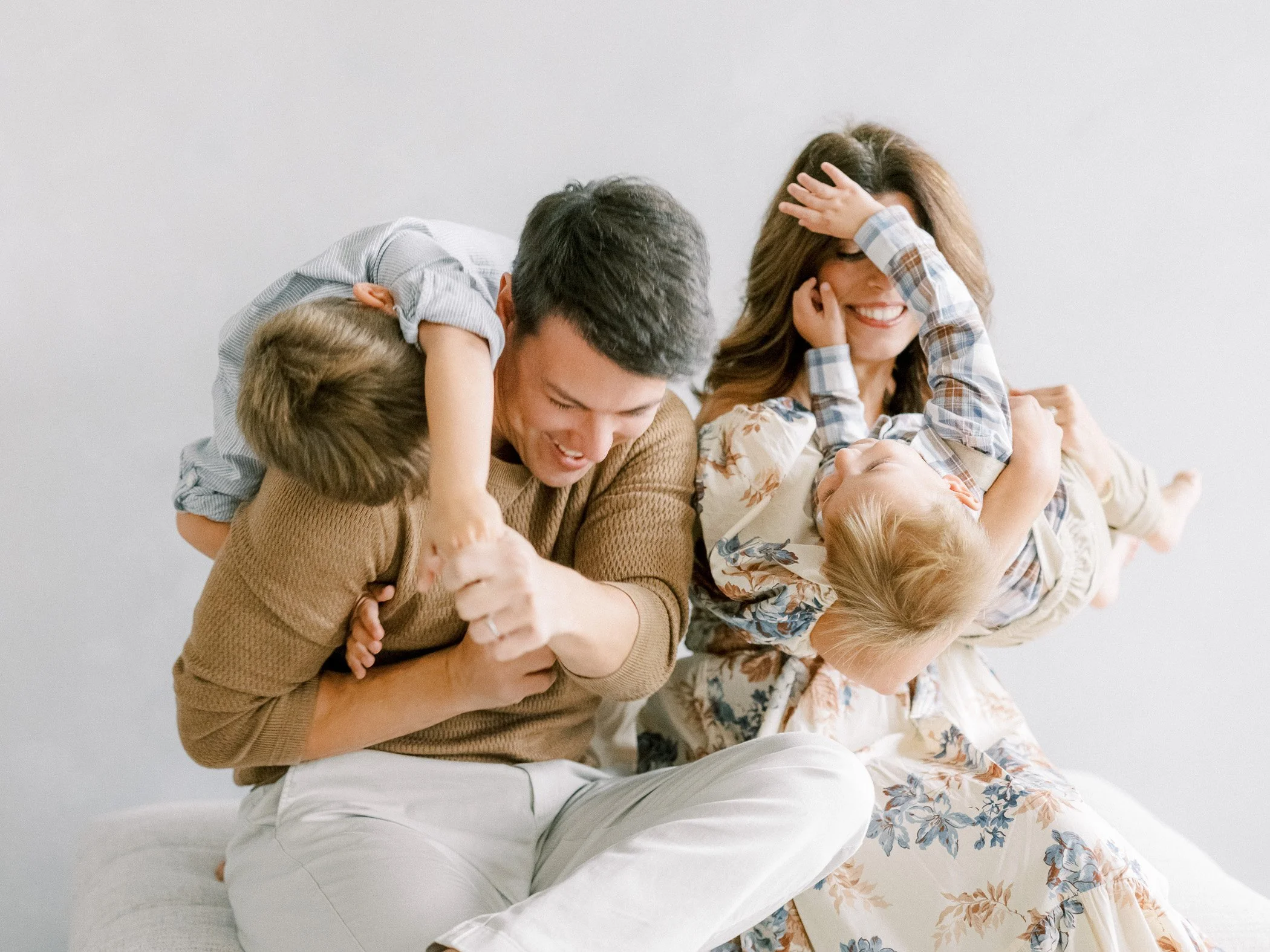 Family of four playing and laughing together on a bed, with children playfully draping over parents.