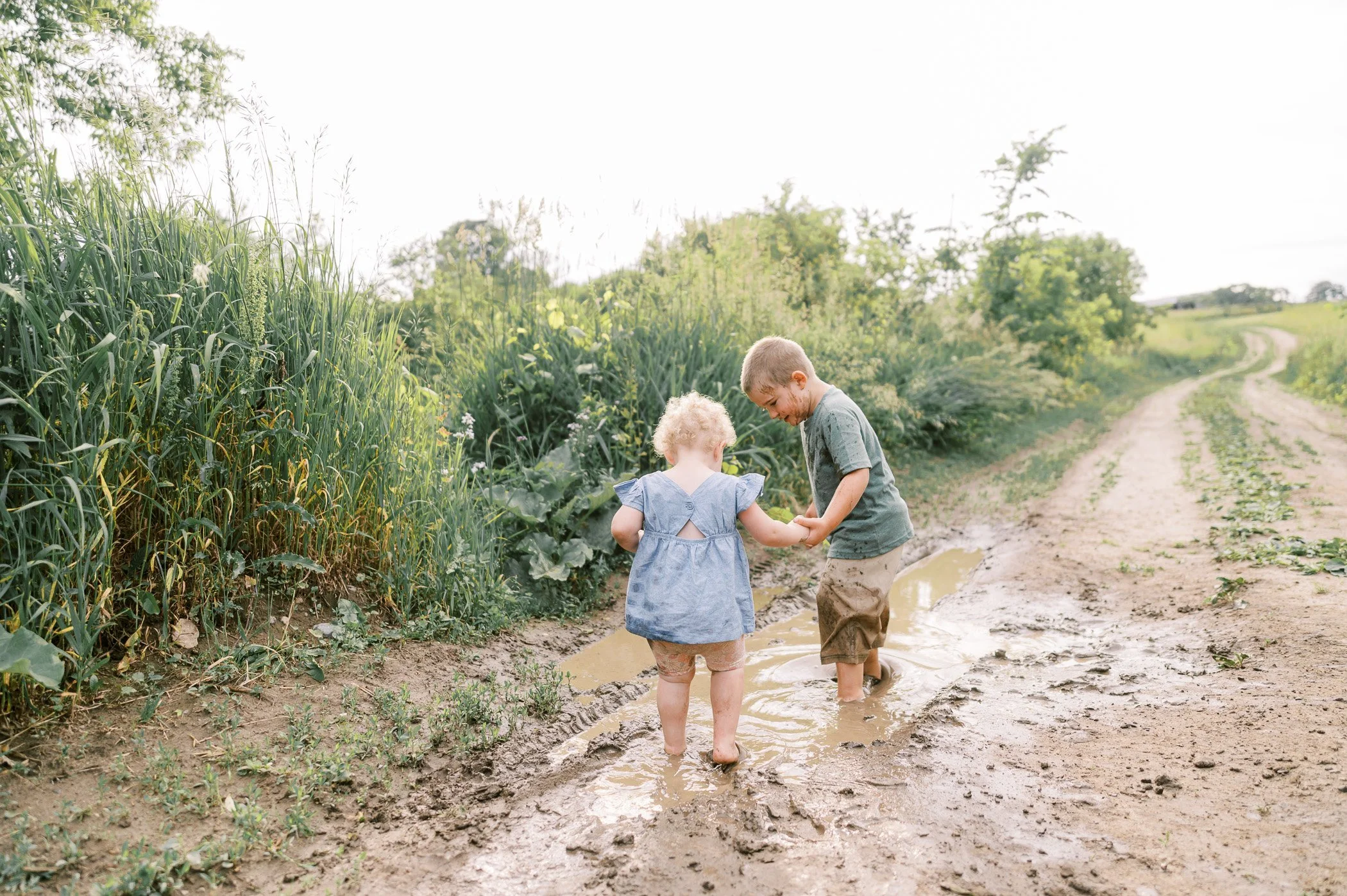 Two children, a boy and a girl, are playing and splashing in a muddy puddle on a dirt path in a rural area surrounded by green plants and trees during daylight.