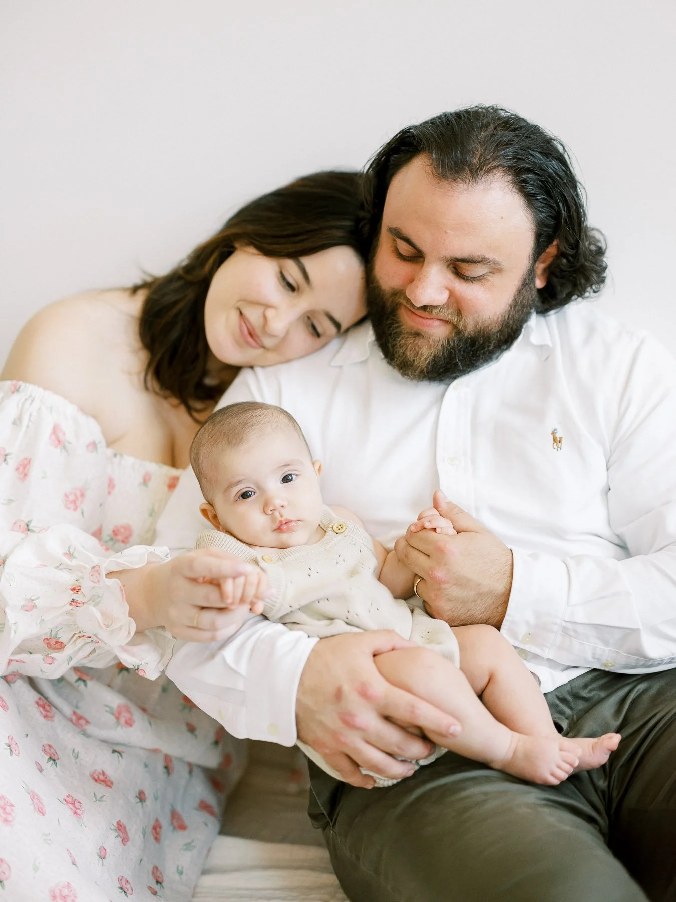 A family of three sitting together, with a mother, father, and their baby, all smiling softly. The mother is leaning her head on the father's shoulder. The baby is sitting on the father's lap, holding onto his arm, with a neutral expression. The sett