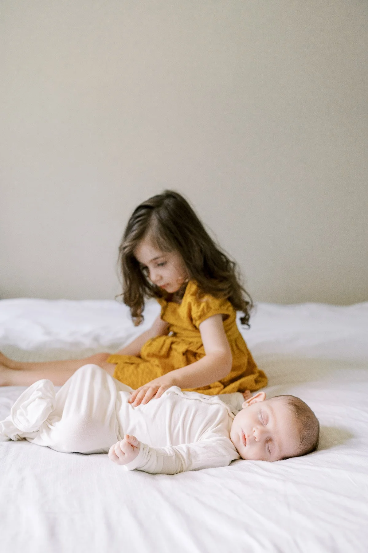 A young girl with brown hair and a yellow dress sitting on a bed, looking down at a sleeping baby in a white onesie.