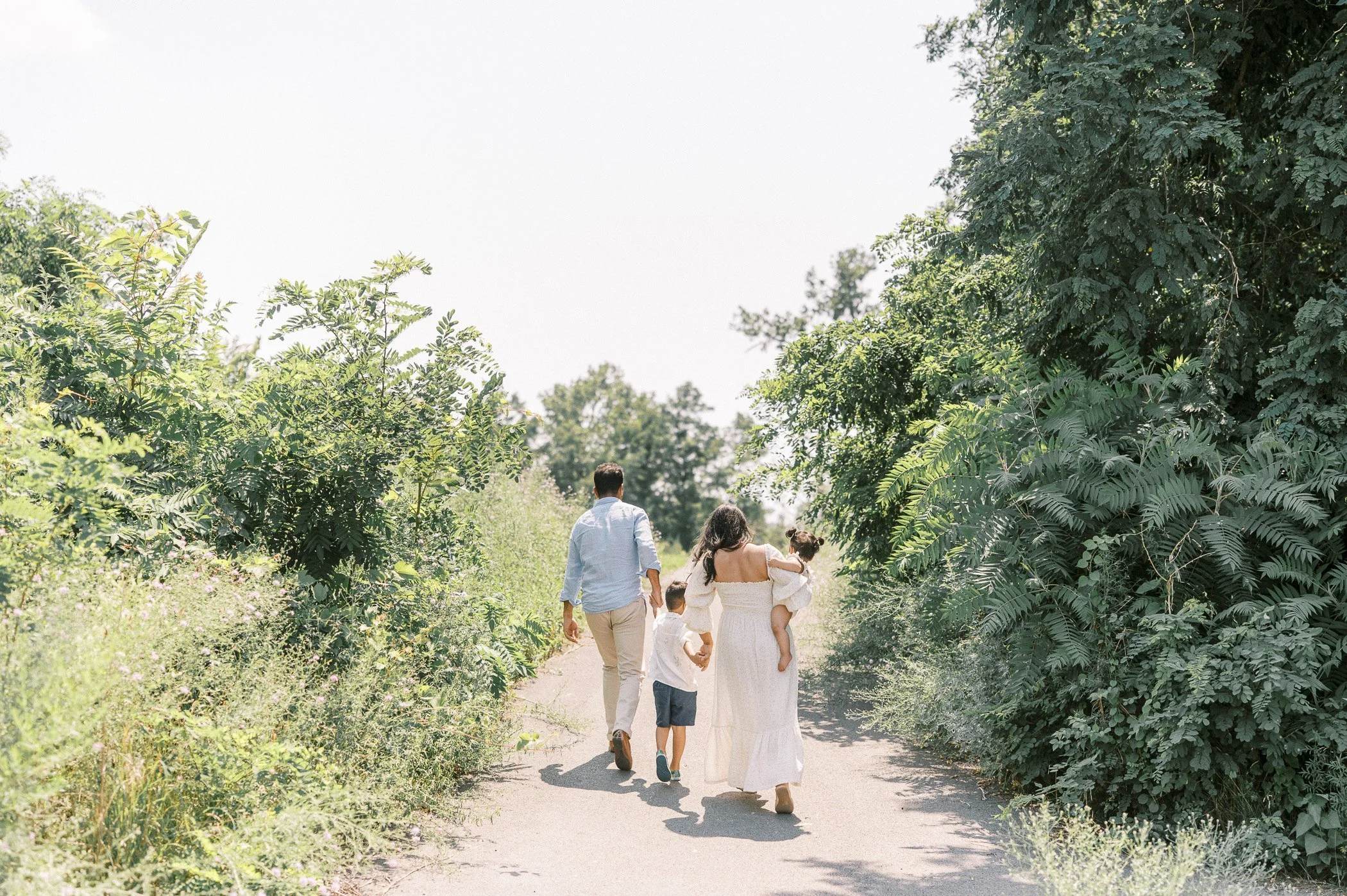 Family of four walking outdoors on a sunny day, surrounded by green trees and bushes, with a dirt path underfoot.
