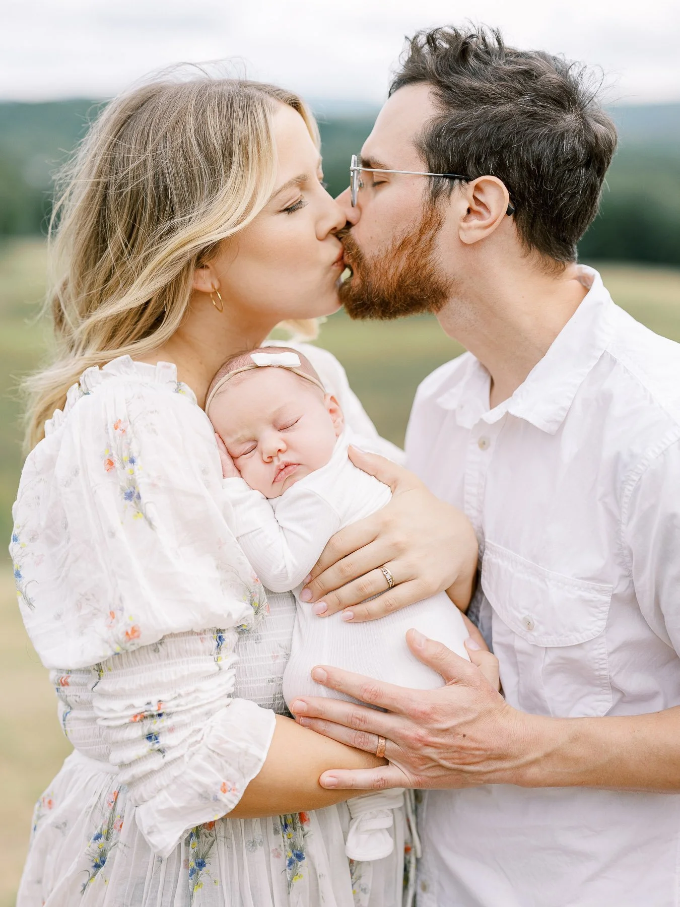 A family of three outdoors, a woman and man kissing while holding their sleeping baby.