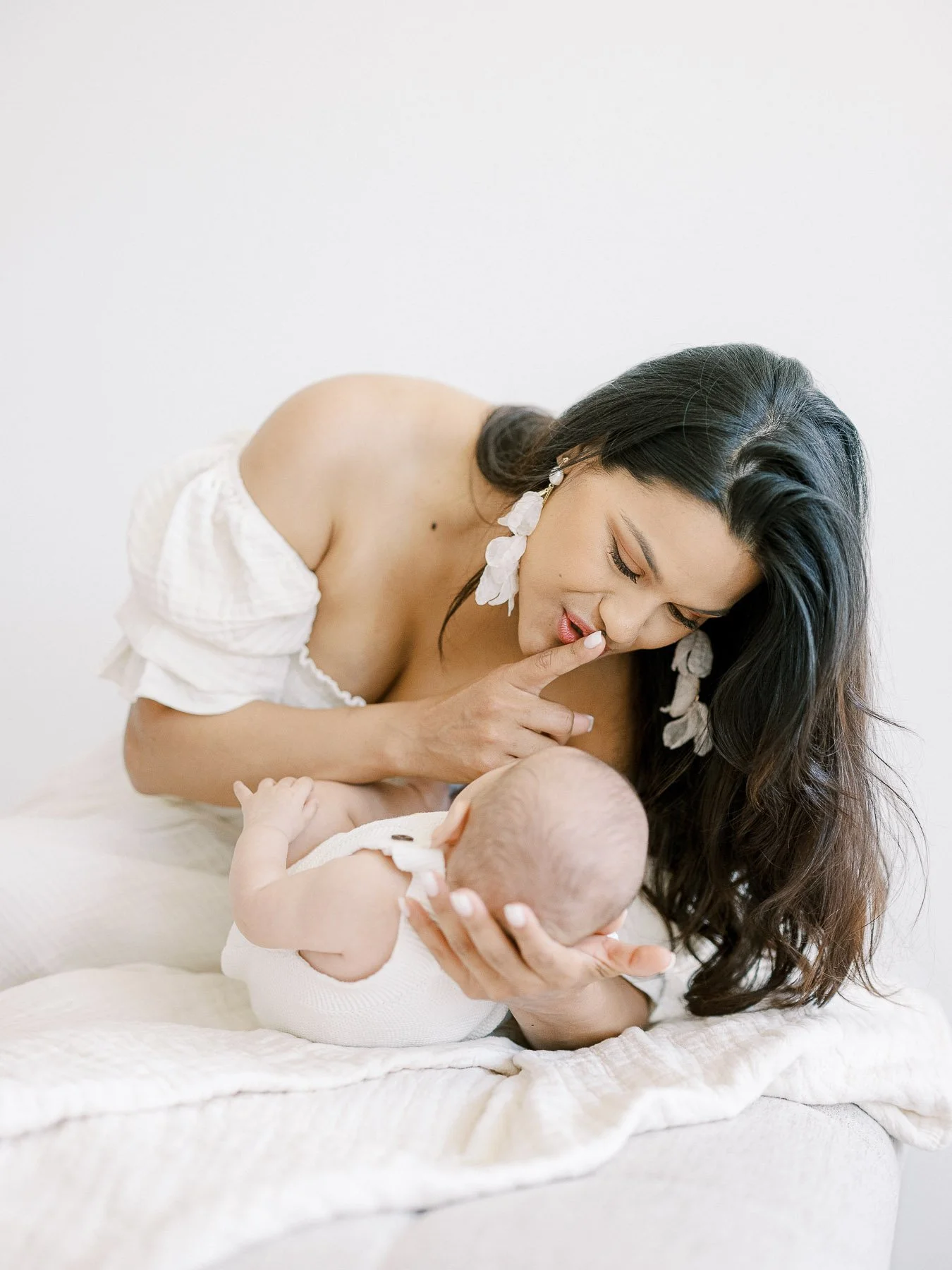A woman with dark hair and floral earrings holding and gently touching a baby's hand, both lying on a white blanket on a bed or couch in a bright, minimal setting.