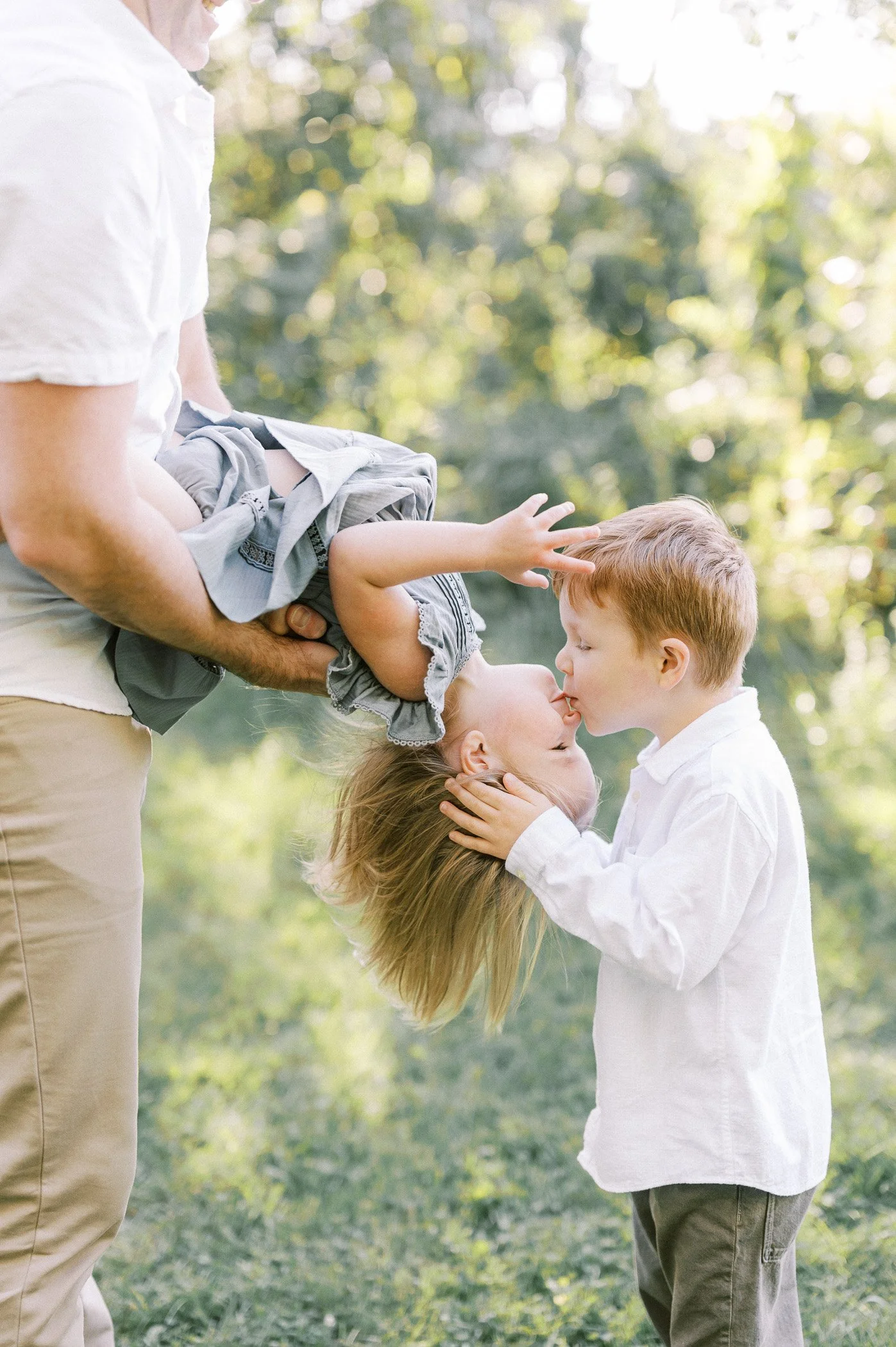 A young girl is being held upside down by a man, while a young boy kisses her on the lips in an outdoor setting with trees and sunlight.