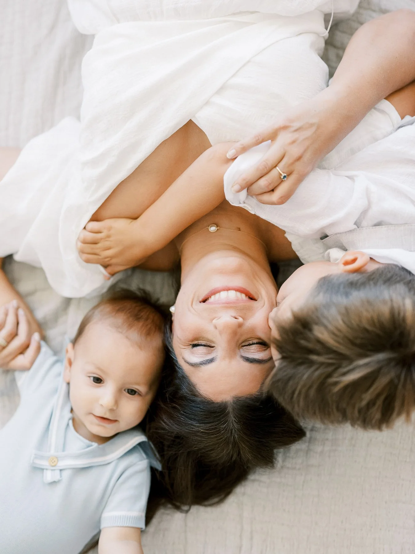 A smiling woman lying on her back on a sofa, surrounded by two children, one on each side, with one kissing her on the cheek and the other lying close to her head. All are in casual clothing.
