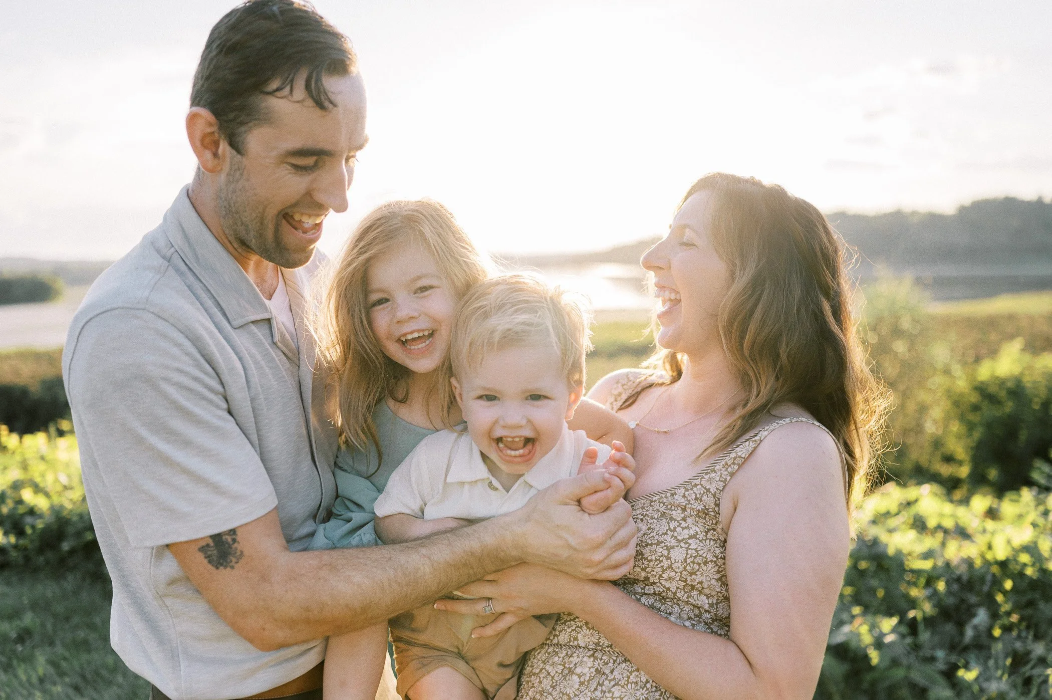 A happy family of four laughing and playing outdoors during sunset with scenic landscape in the background.