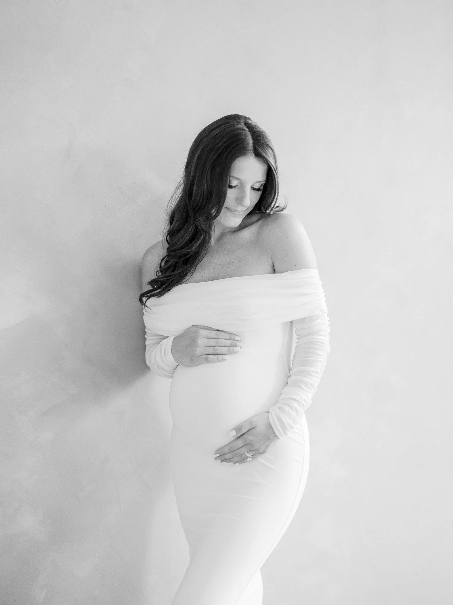 Black and white photo of a pregnant woman in a form-fitting dress, gently holding her baby bump and looking down.