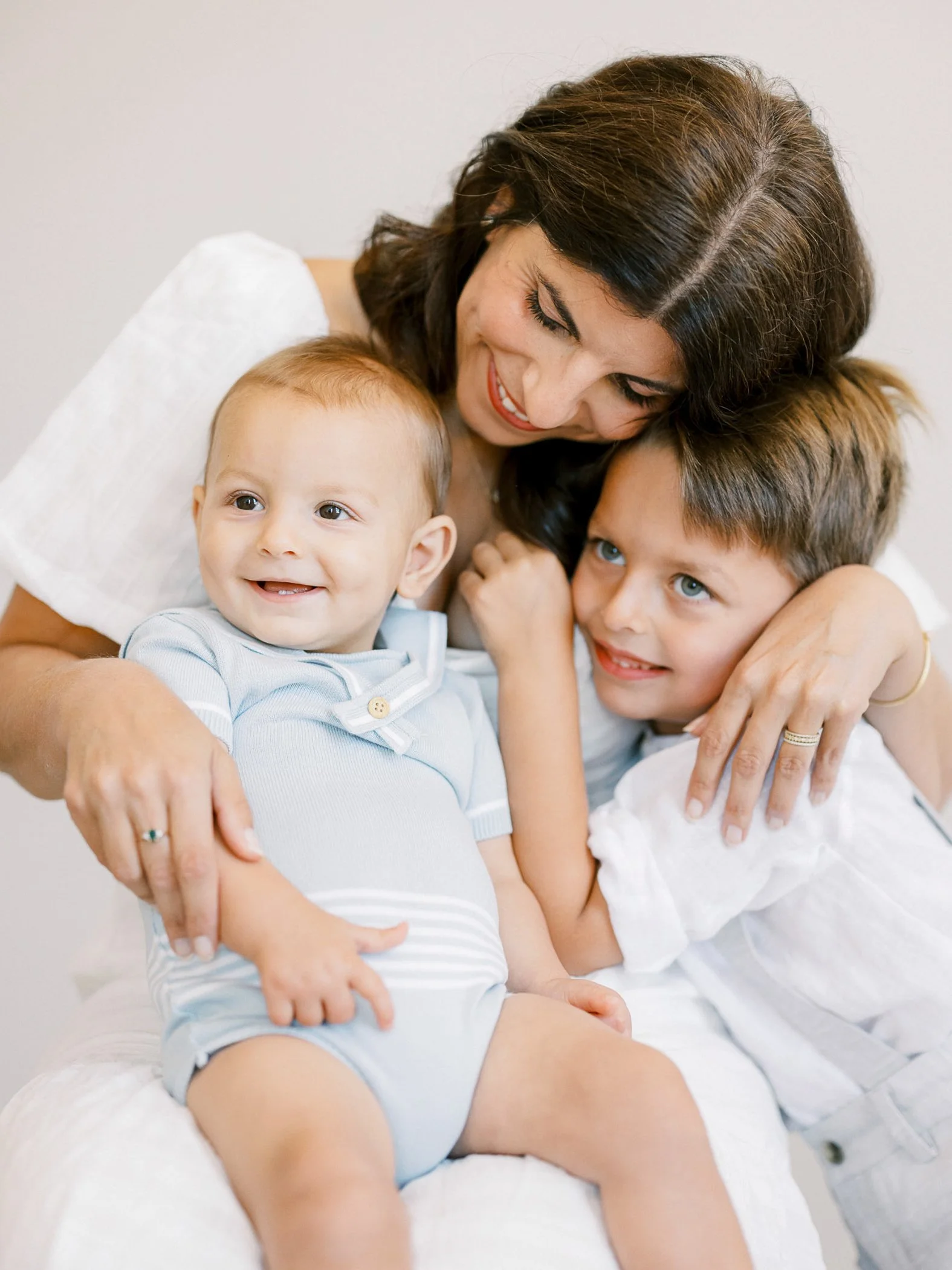 A woman with dark hair hugging two young boys, one with brown hair and the other with light hair, all smiling and close together.
