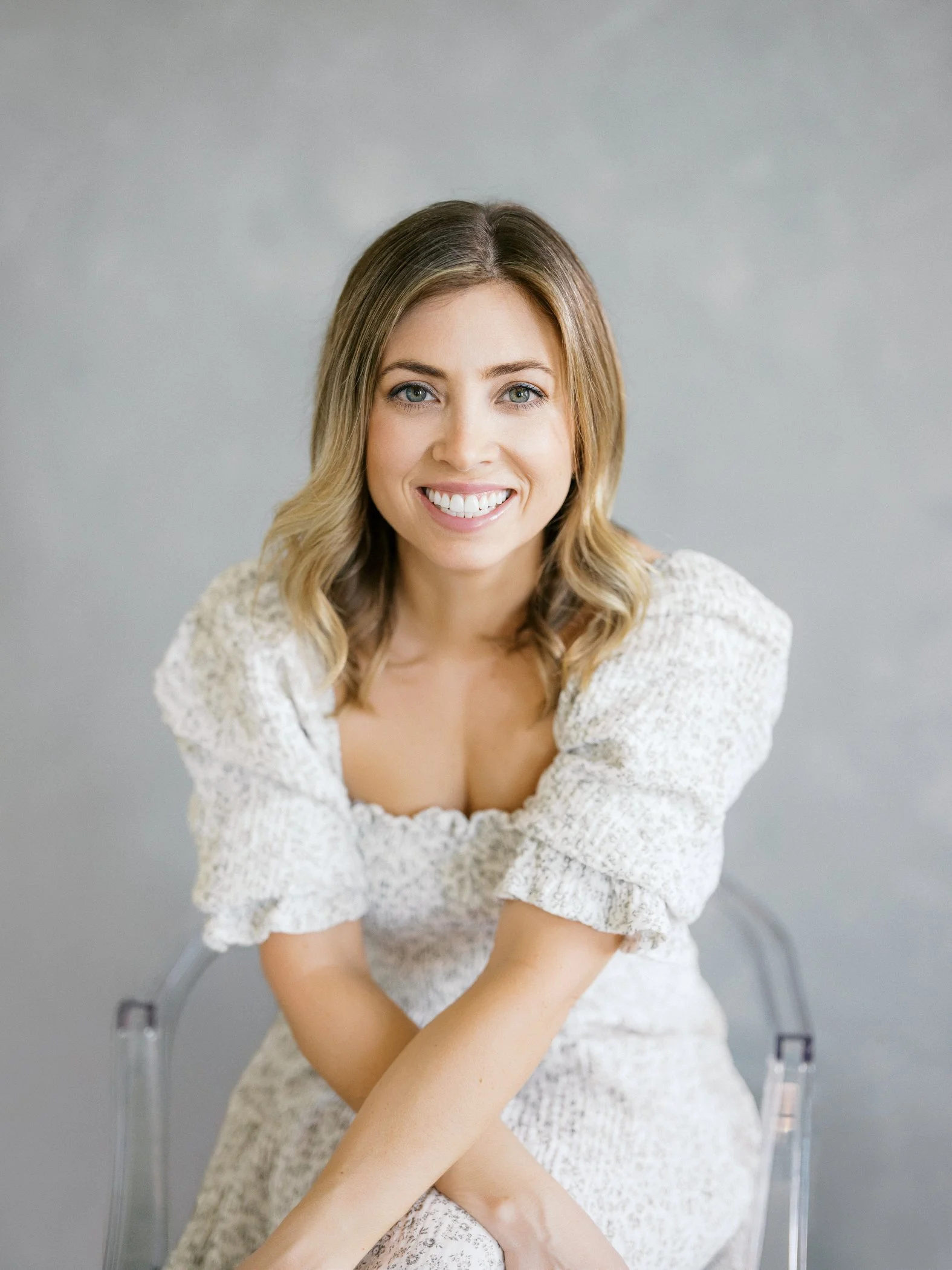 A woman with shoulder-length blonde hair and blue eyes, smiling, wearing a white dress with puffed sleeves, sitting on a transparent chair against a gray background.