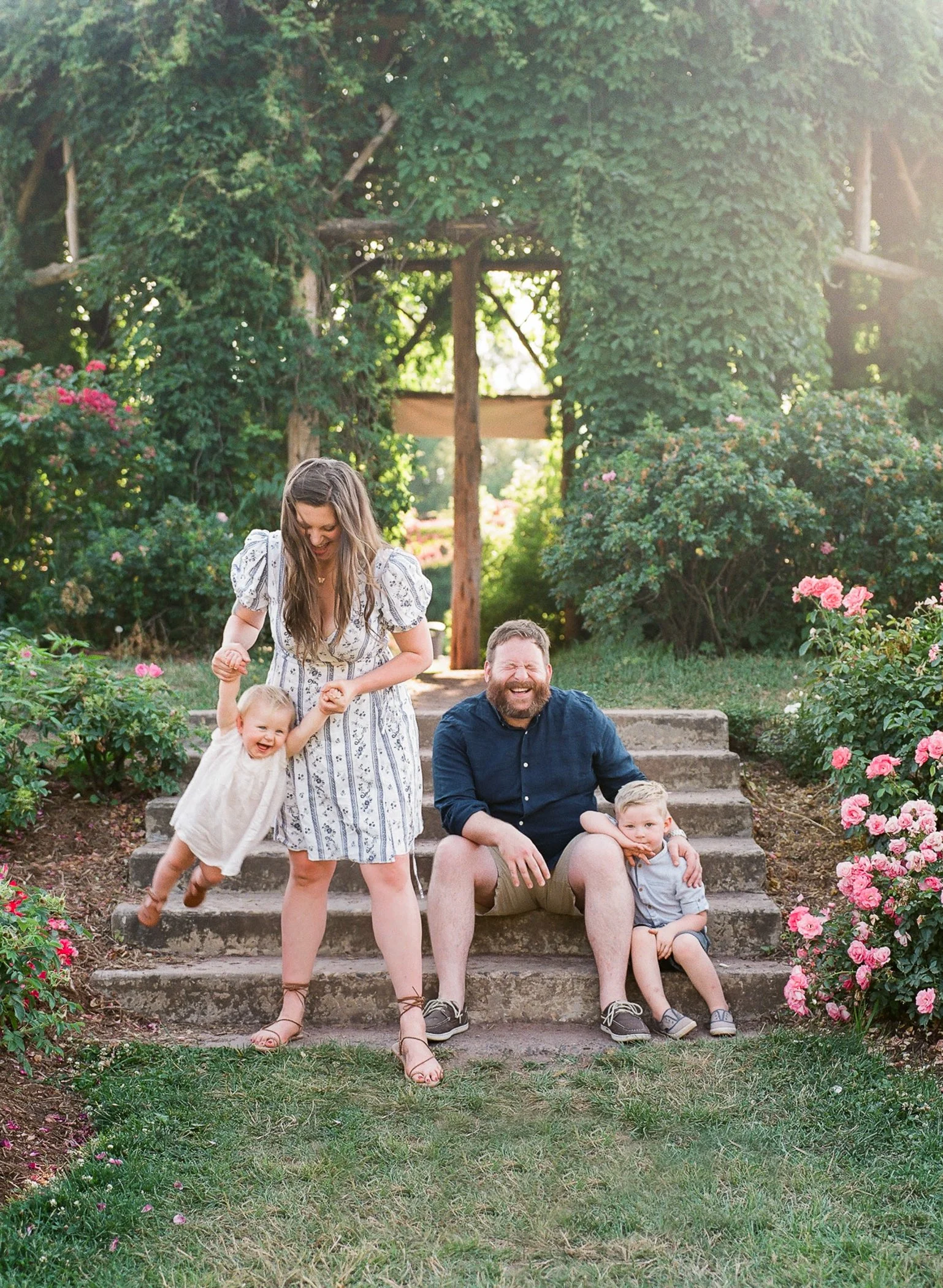 A family of four outdoors on stone steps, surrounded by pink flowers and lush green trees, smiling and enjoying a sunny day.