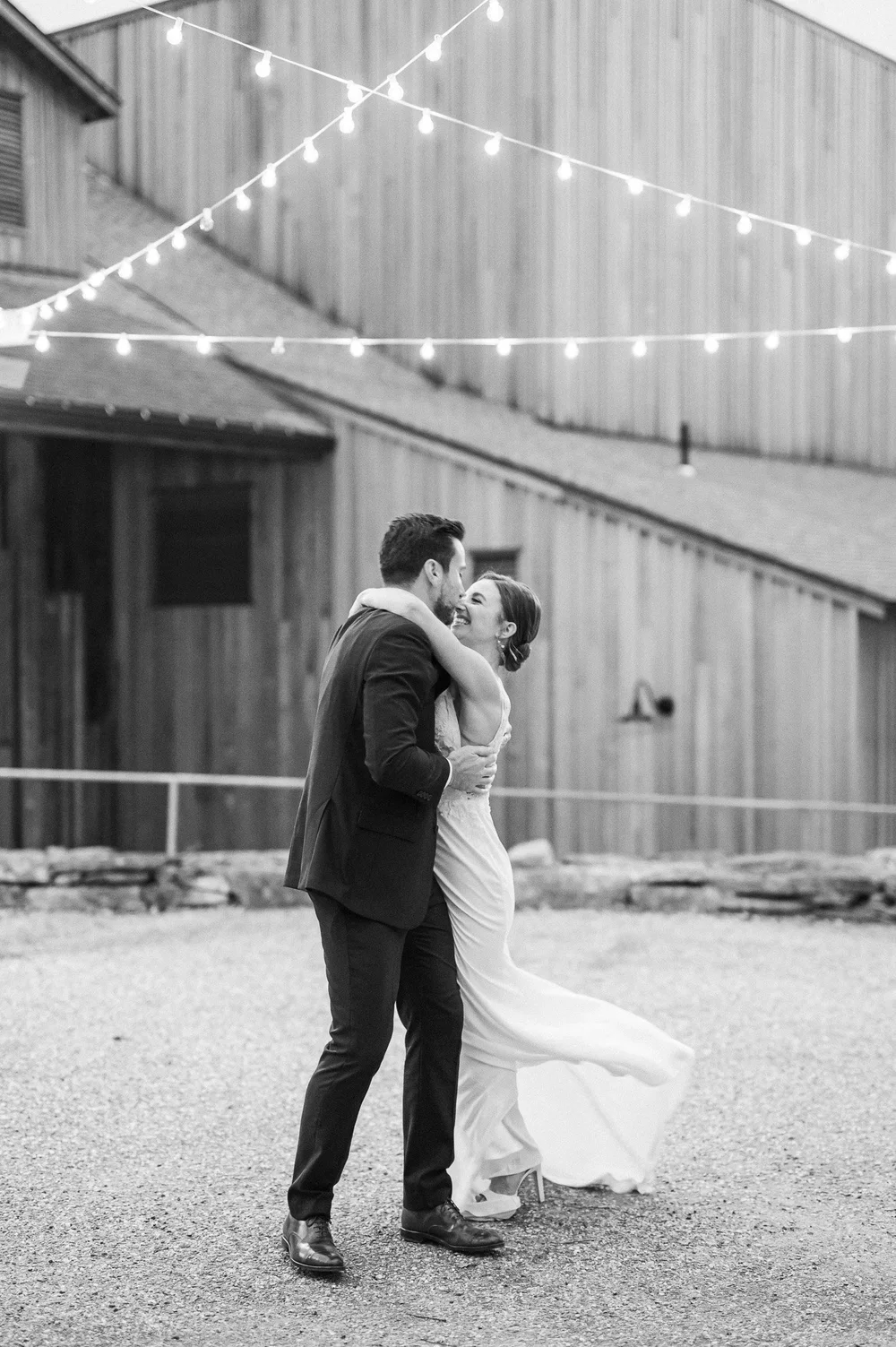 A black-and-white photo of a couple dancing outdoors at night, illuminated by string lights, with a rustic wooden building in the background.