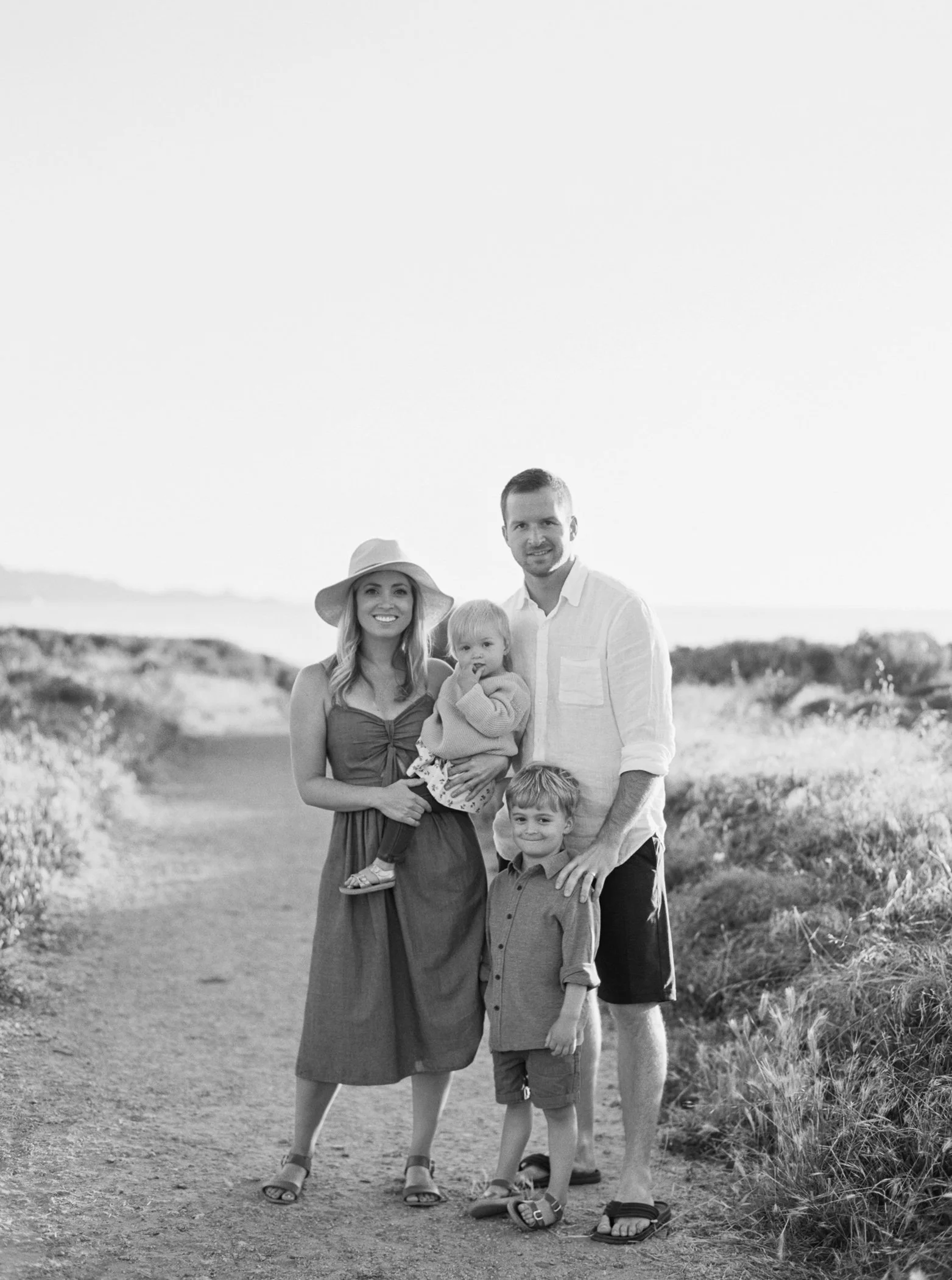Family of four standing outdoors on a dirt path with bushes and sky in the background. Woman wearing a hat and dress, holding a young girl. Man in a white shirt and shorts, standing next to a young boy. Black and white photo.
