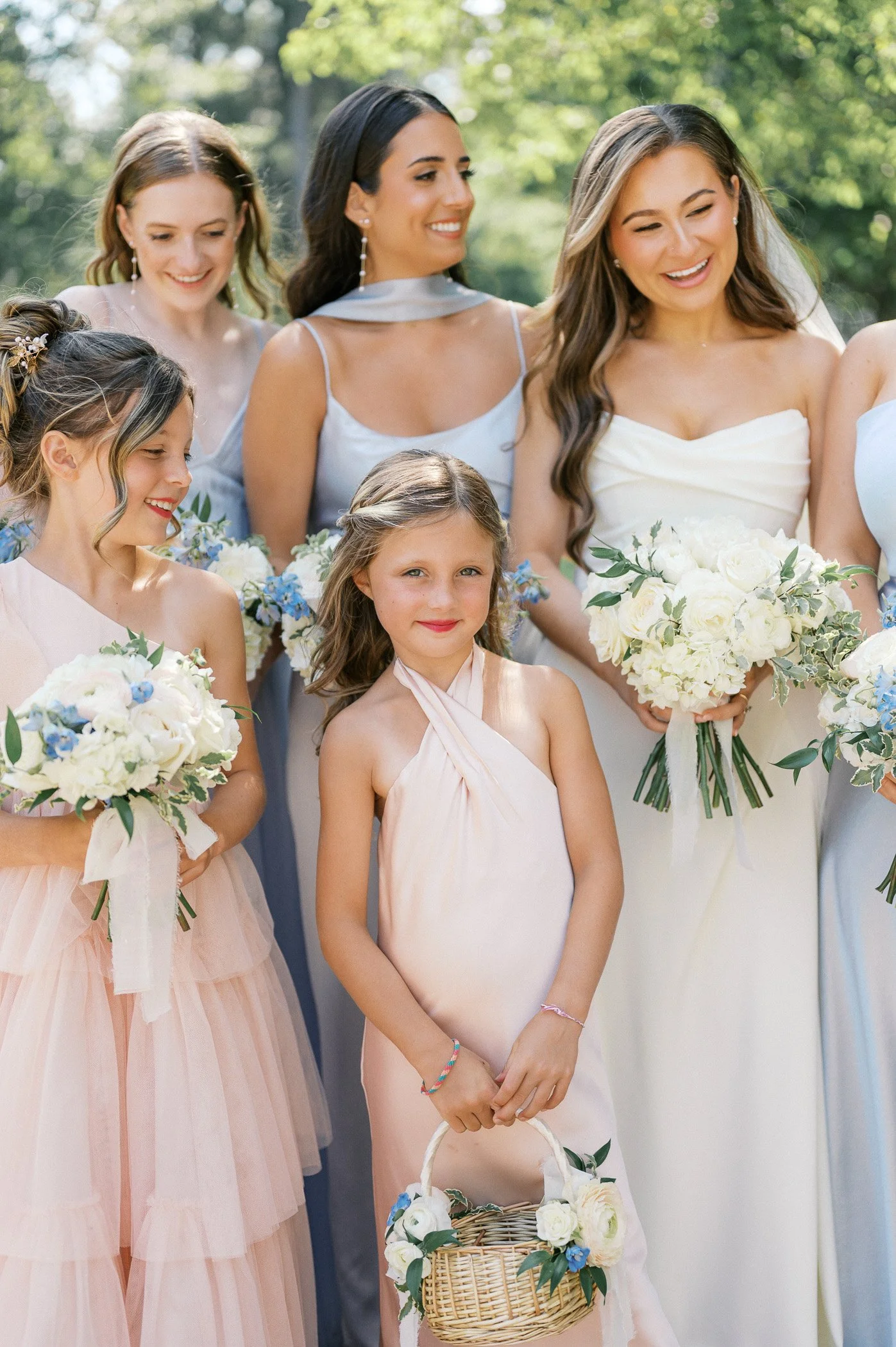 Group of women and children dressed in pastel and white dresses, holding bouquets of white and blue flowers, standing outdoors in a garden setting for a wedding celebration.